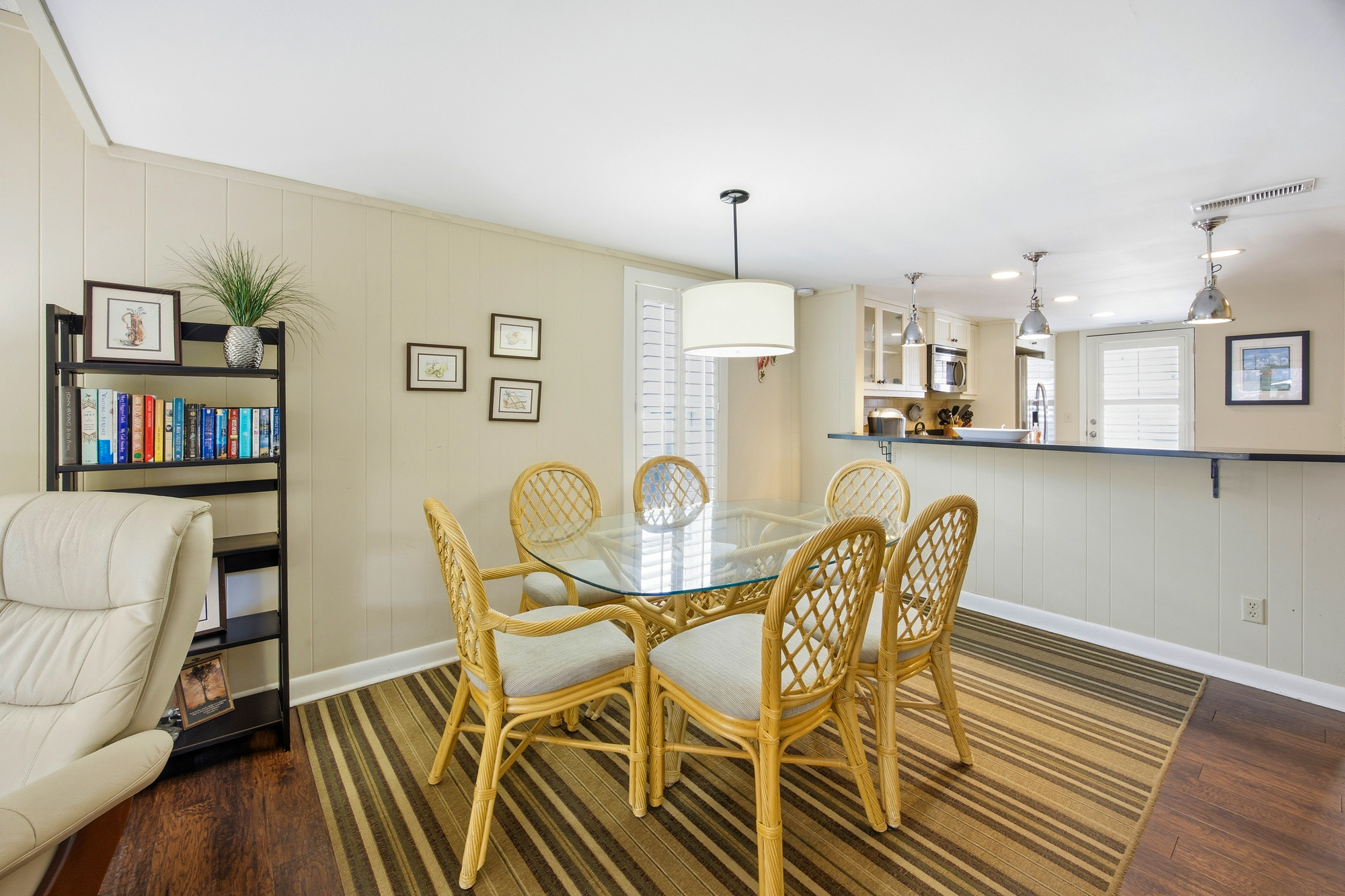 Rattan chairs and a glass table create a breezy dining nook perfect for long breakfasts. Golden tones from the wood floor and cream walls glow under the drum pendant, making this open layout feel peaceful and bright.