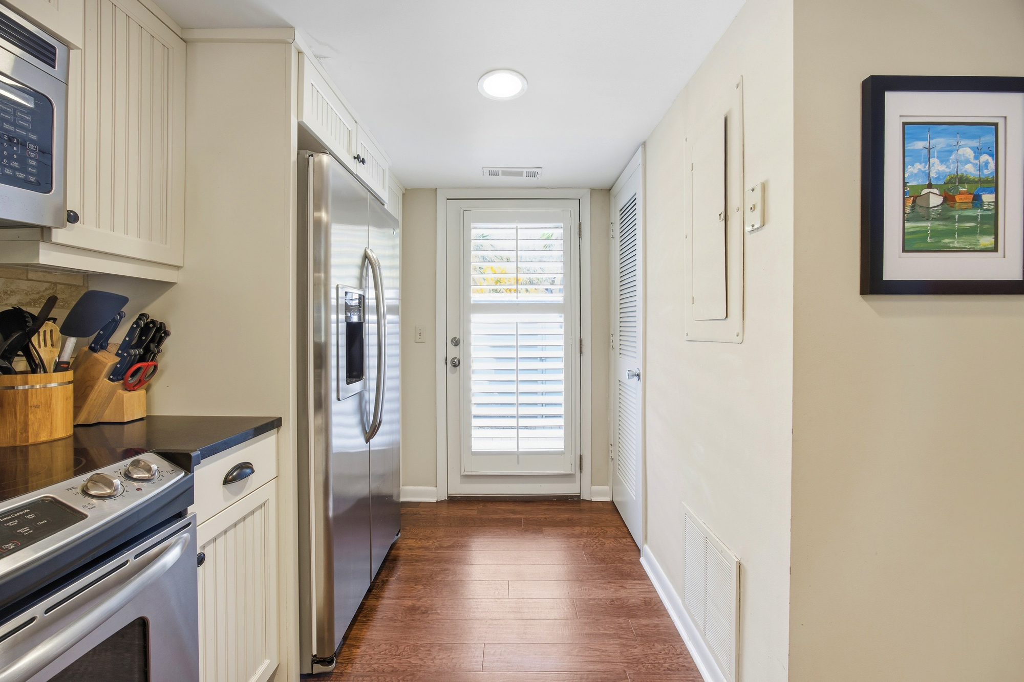 Stainless steel appliances gleam in this efficient kitchen, where rich wood floors lead your eye toward a sun-washed door. Shutter-filtered rays brighten the clean white cabinetry and nautical art, creating an airy spot for morning coffee.
