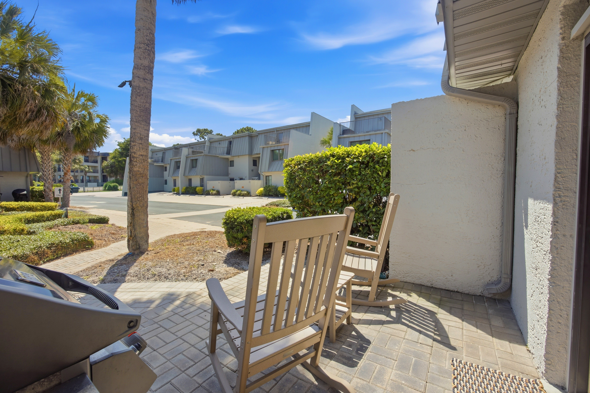 Wooden rocking chairs wait on the paved patio, offering a quiet corner for morning coffee and conversation. Palm trees sway under a bright azure expanse, casting long shadows across this peaceful outdoor nook.