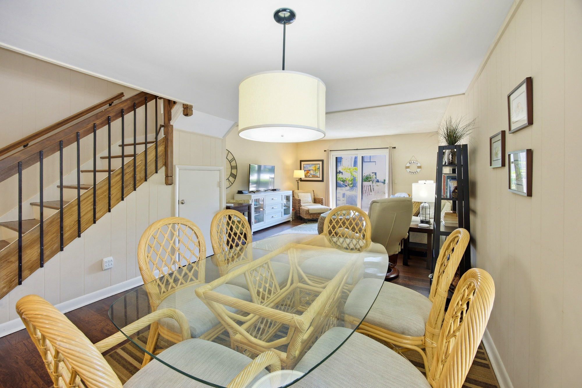 Honey-hued wicker chairs and a sleek glass table anchor this airy dining space, where a drum pendant casts a gentle glow. Rich wood floors and cream paneled walls lead your eye toward the sun-drenched patio just beyond the living room.