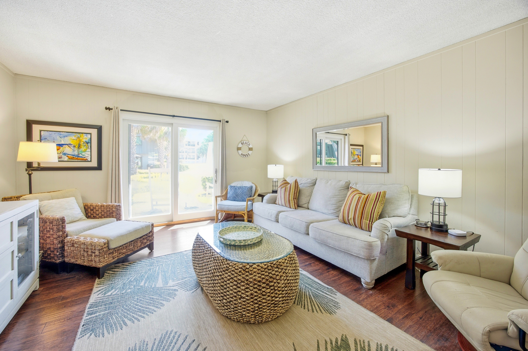 Placid neutral tones and organic textures create a serene living area where afternoon brightness pours through the glass. Thick seagrass accents and a palm-leaf rug bring a touch of the outdoors into this airy, comfortable common room.