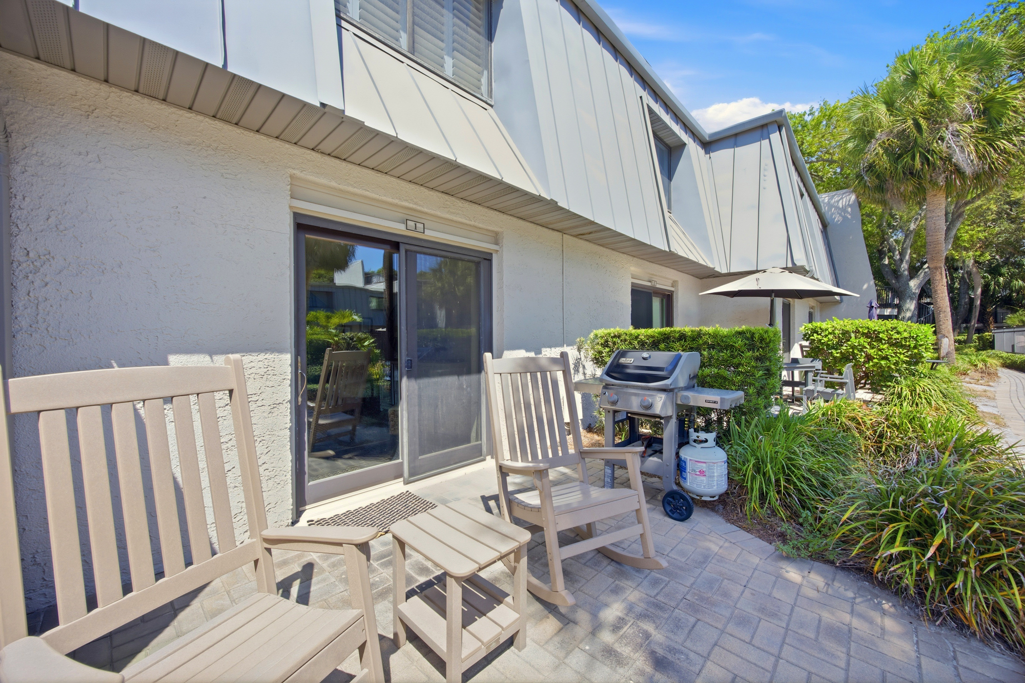 Weathered rocking chairs rest on a sun-drenched stone patio, offering a quiet corner for morning coffee. Palm trees sway over the gas grill and lush greenery, creating a private outdoor nook for slow afternoons and evening cookouts.