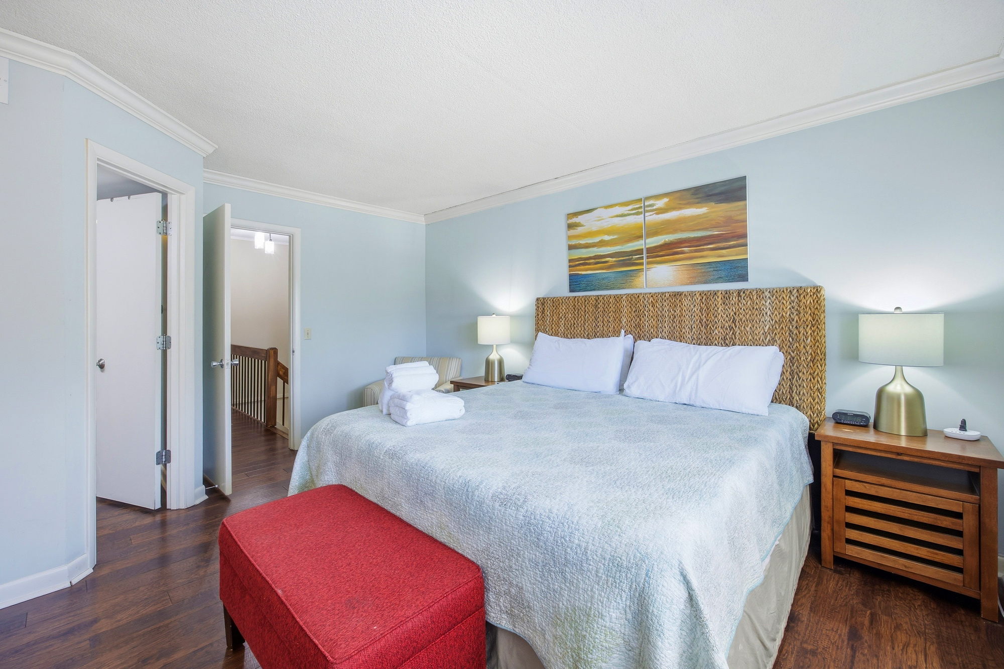 Woven textures and soft blue walls create a serene coastal mood in this spacious bedroom. A plush red ottoman adds a pop of rich color against the dark wood floors, while glowing lamps and sunset art make for a cozy, peaceful atmosphere.