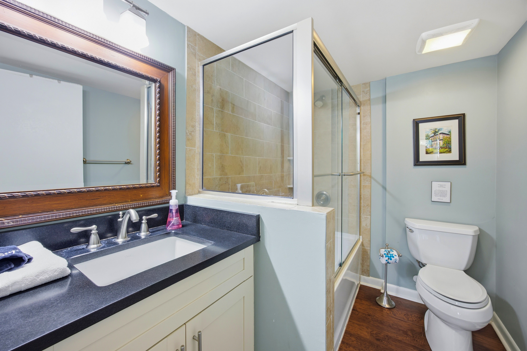Polished granite and rich wood floors anchor this tidy bathroom, where soft blue walls create a calming atmosphere. A framed vanity mirror and glass-enclosed shower offer a bright, functional space for your morning routine.