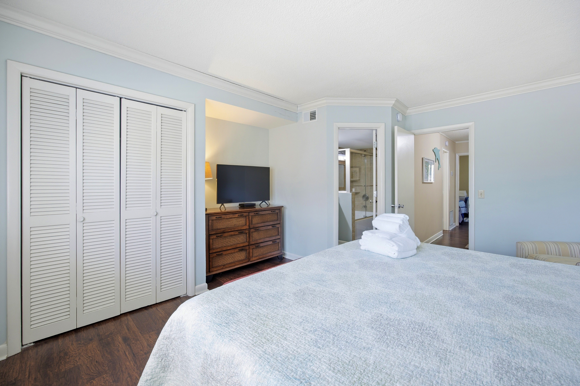 Pale blue walls create a calming backdrop for this restful bedroom, where dark wood floors meet coastal textures. Soft linens and louvered closet doors invite you to slow down and settle in for a quiet night.
