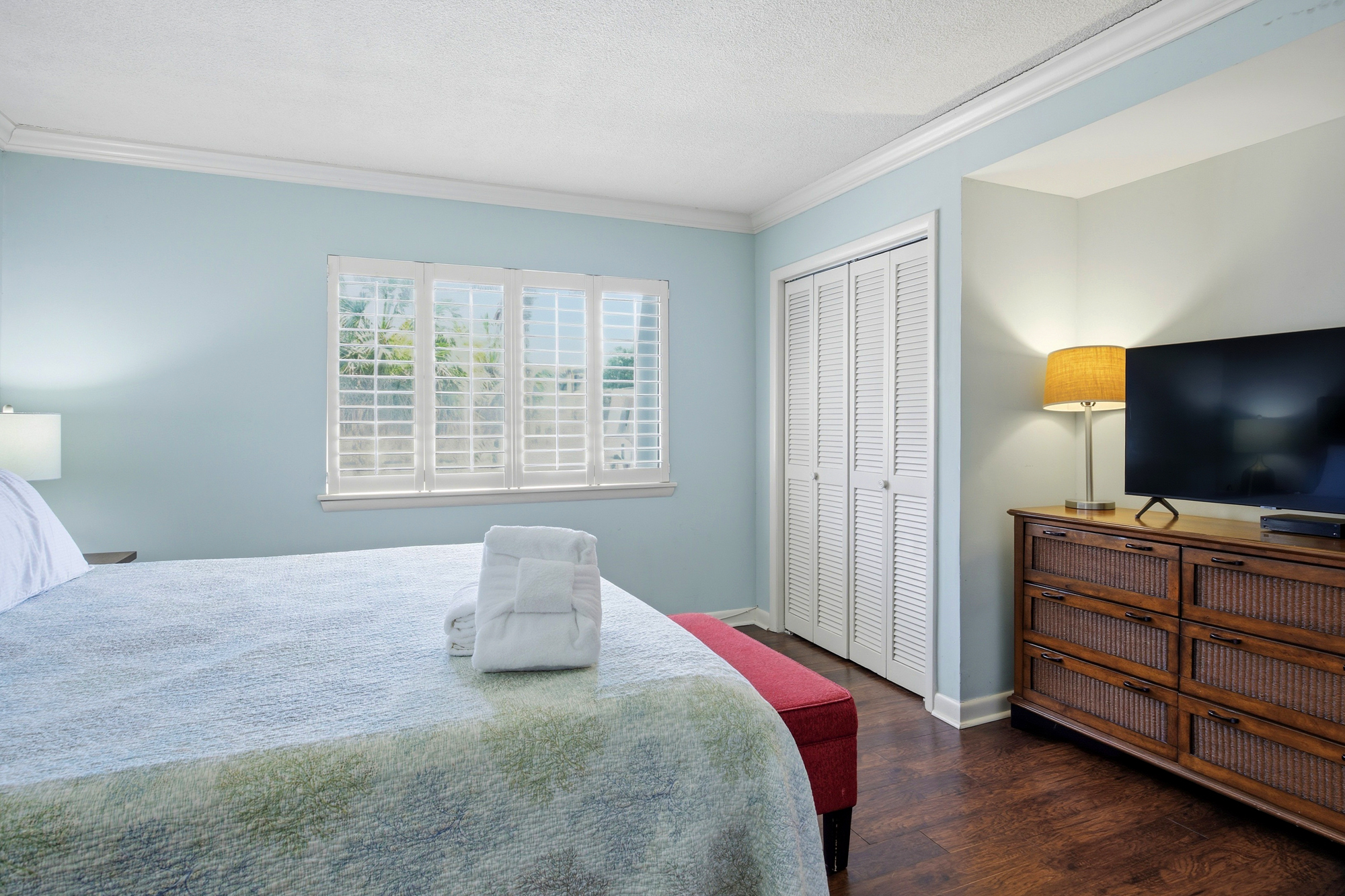 Sky-blue walls and rich wood floors create a serene backdrop for this quiet bedroom. Natural light filters through classic white shutters, casting a gentle glow over the plush linens and woven textures.