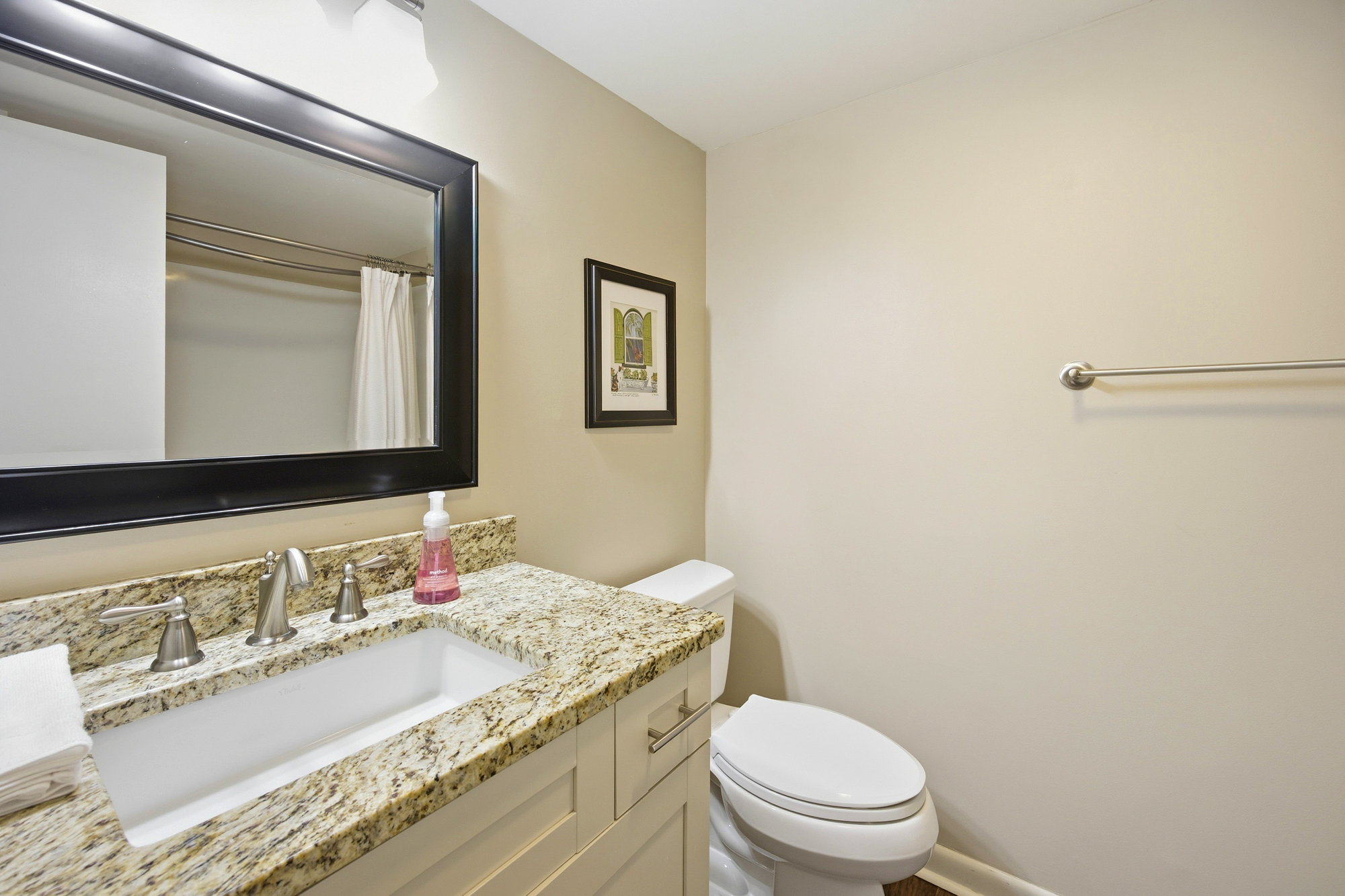Granite counters add a speckled texture to this bright bathroom, where a large framed mirror reflects the clean space. Brushed nickel fixtures and neutral walls create a calm, tidy atmosphere for your morning routine.
