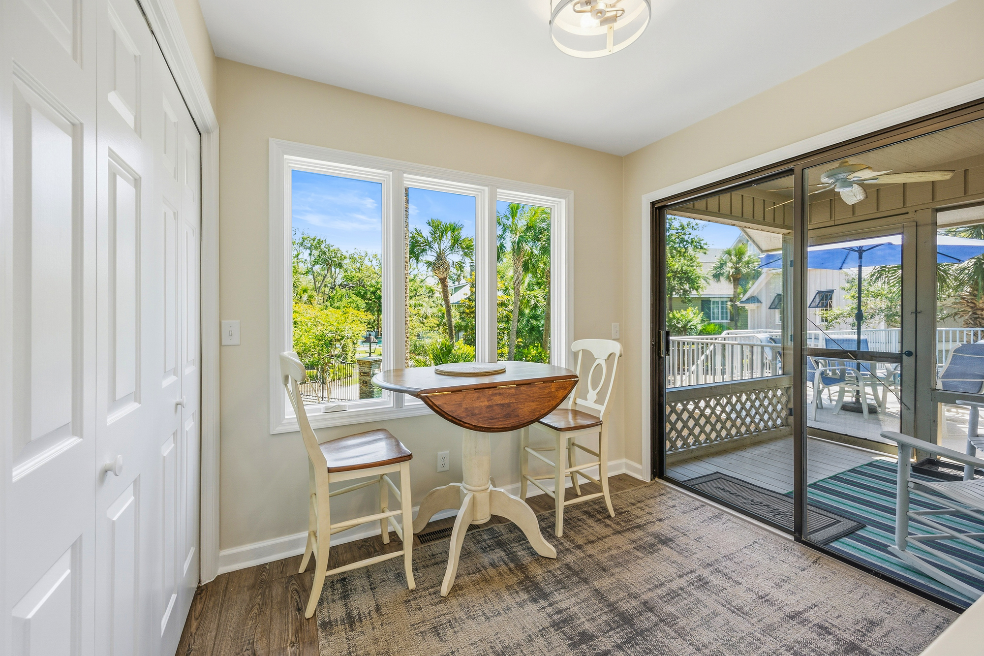 Polished wood textures and creamy white finishes create a soft breakfast nook where morning rays pour through wide glass panes. Sliding doors open to a breezy deck, blending indoor comfort with the swaying palms of the garden view.