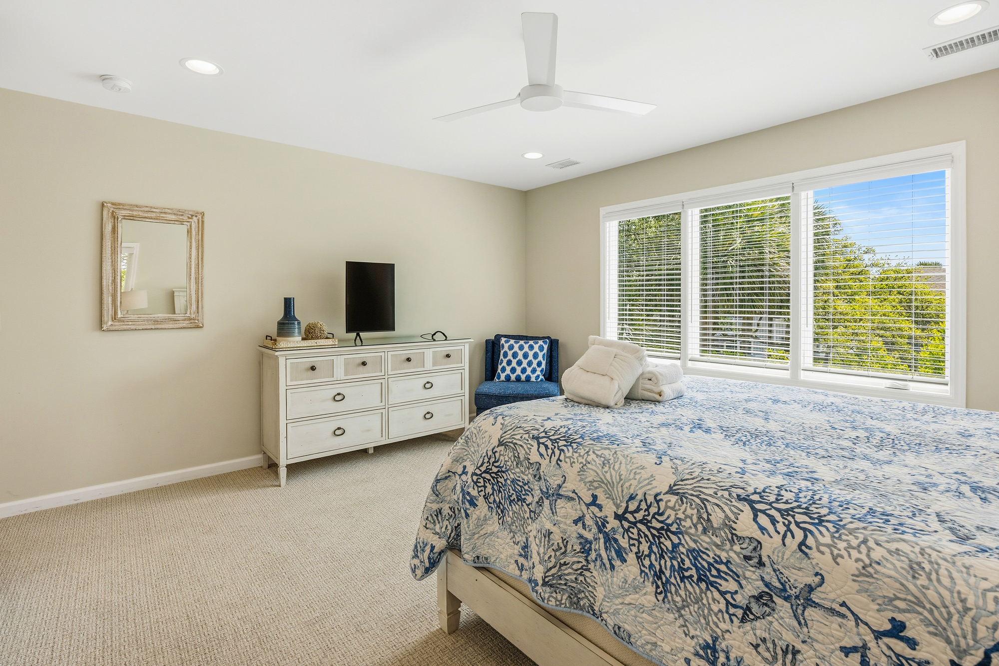 Coastal blues and sandy tones create a serene atmosphere in this airy bedroom. Large windows invite a bright, natural glow to dance across the coral-patterned bedding, offering a peaceful space to recharge after a day by the sea.