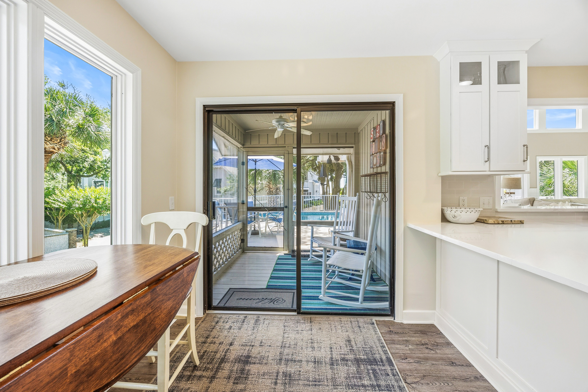 Rich walnut wood tones meet airy white cabinetry in this bright kitchen and dining nook. Sliding glass doors pull your gaze toward a screened porch where rocking chairs wait for a slow morning coffee in the coastal breeze.