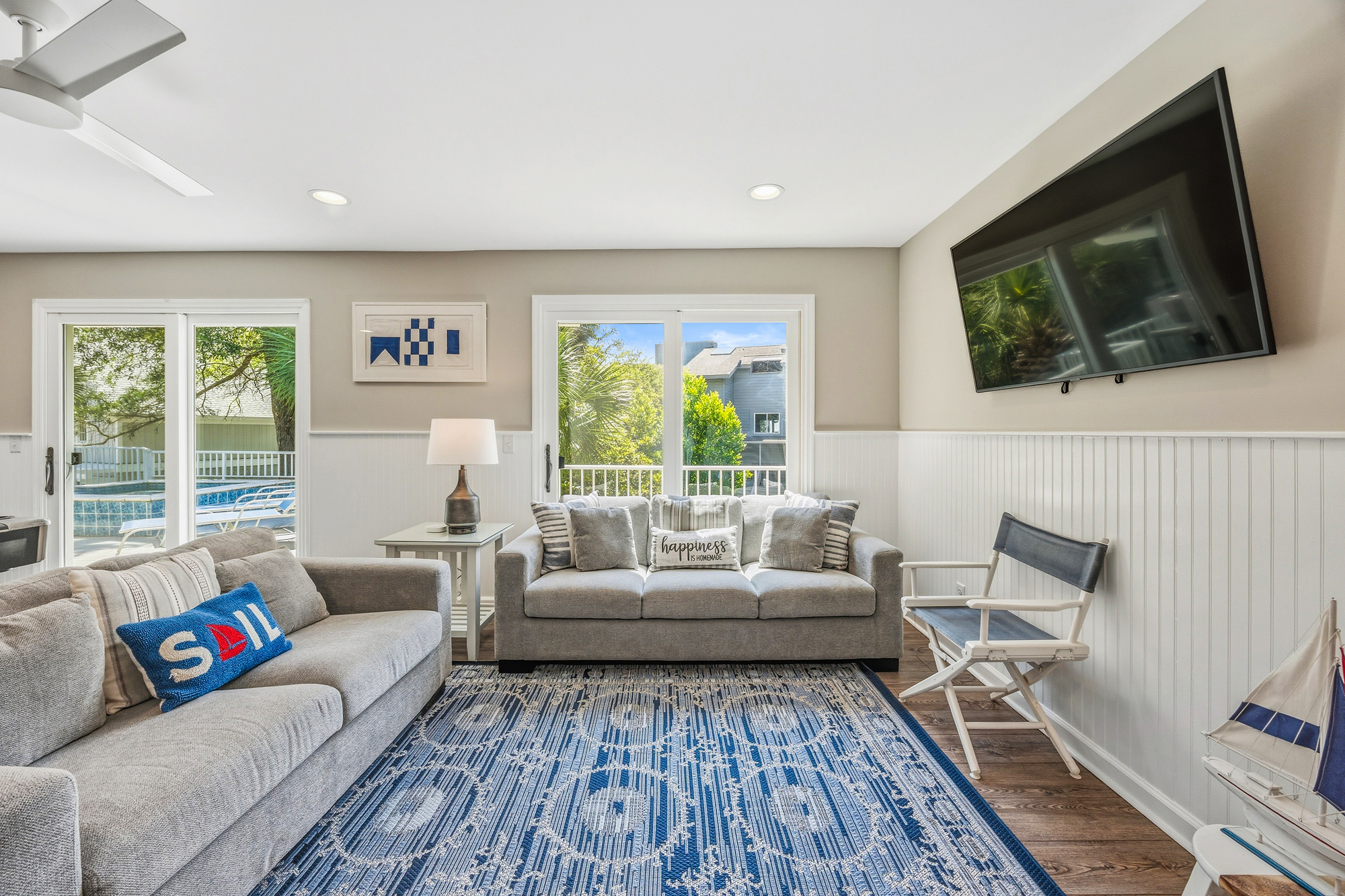 White wainscoting and nautical accents create a breezy atmosphere in this sun-drenched living area. Plush seating pairs with a patterned rug to offer a comfortable spot for movie nights or watching the palms sway through the glass doors.