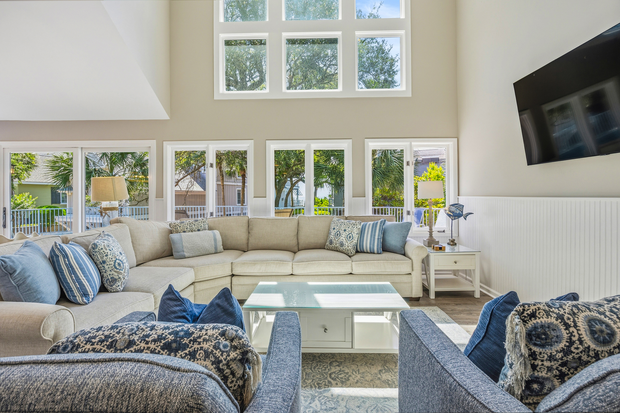 Plush denim tones and creamy fabrics fill this airy living room, where towering windows invite the afternoon glow to dance across the glass coffee table. This soaring space blends soft textures with leafy views for a perfect social hub.