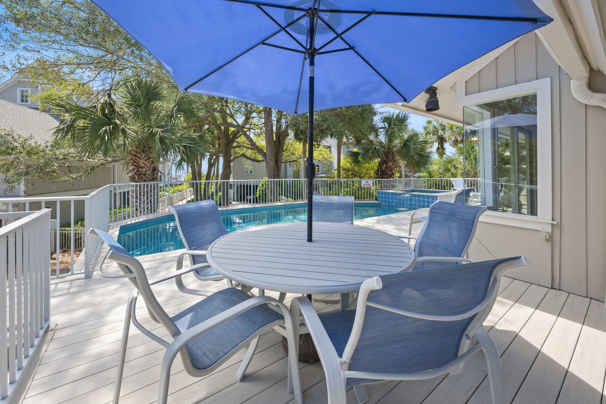 Azure umbrellas cast a cool glow over the weather-worn deck, creating a breezy spot for morning coffee by the sparkling pool. Palm fronds sway in the distance, framing this quiet corner for alfresco dining and afternoon lounging.