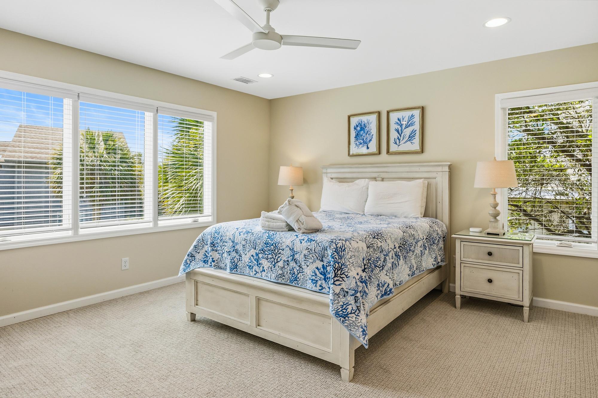 Coral patterns and ocean blue hues bring a calm energy to this airy bedroom, where soft natural light filters through wide windows. Distressed wood furniture and plush carpeting create a soothing space for a restful night by the sea.