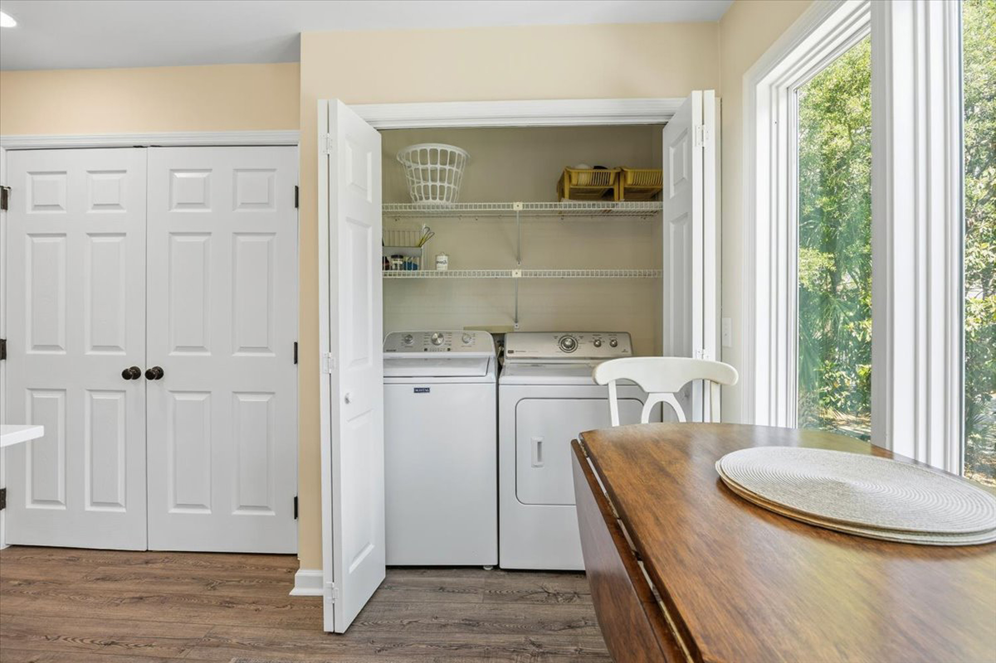 Convenient laundry facilities are tucked behind folding doors, keeping chores simple and out of sight. Natural light pours in from the garden window, illuminating the wood table and creating a bright, efficient utility corner.