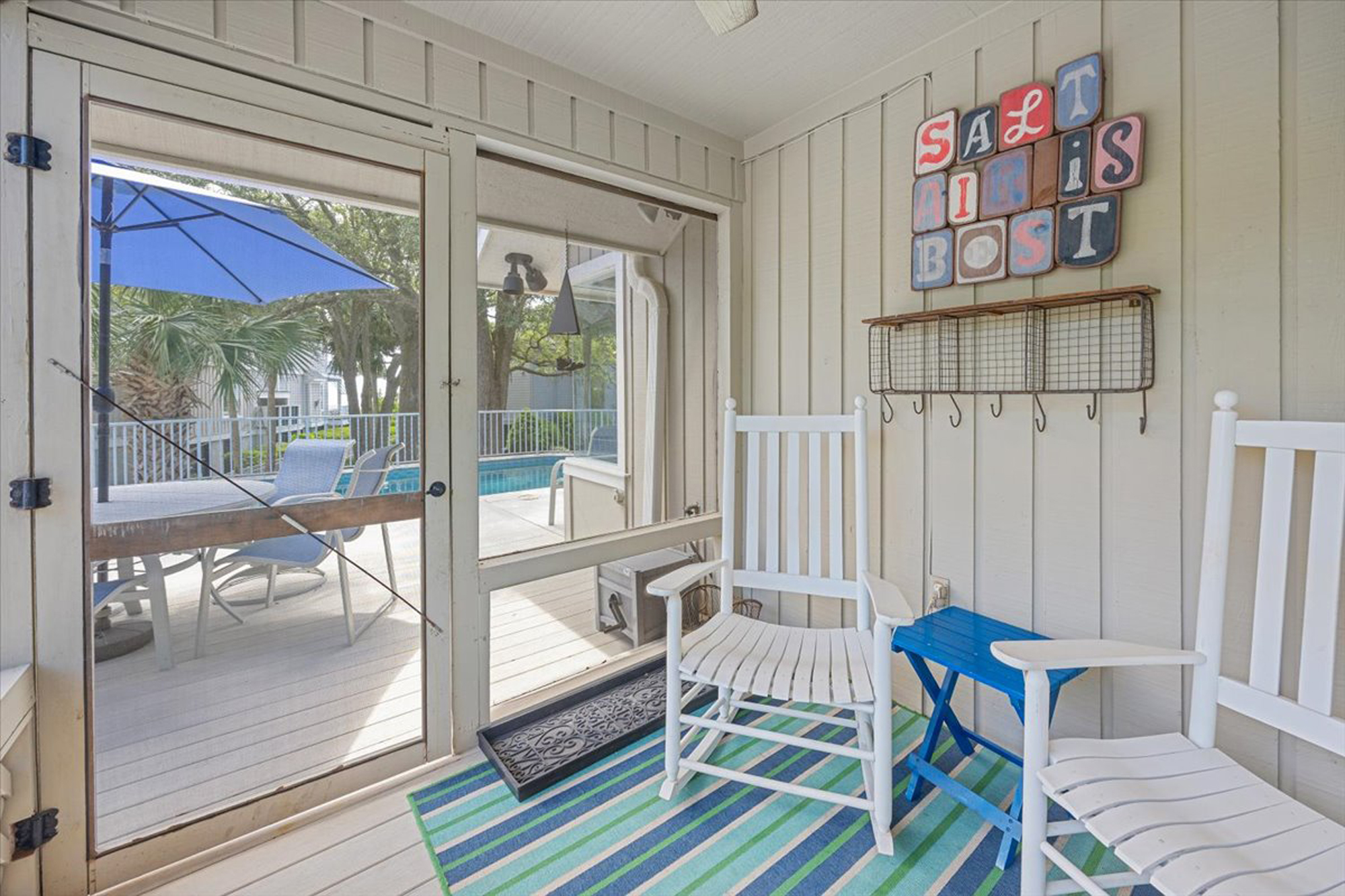 Sun-bleached rocking chairs rest upon a striped rug, offering a quiet corner to watch the pool through the screen. Shiplap walls and whimsical art create a breezy, casual atmosphere for slow mornings and cool evening drinks.
