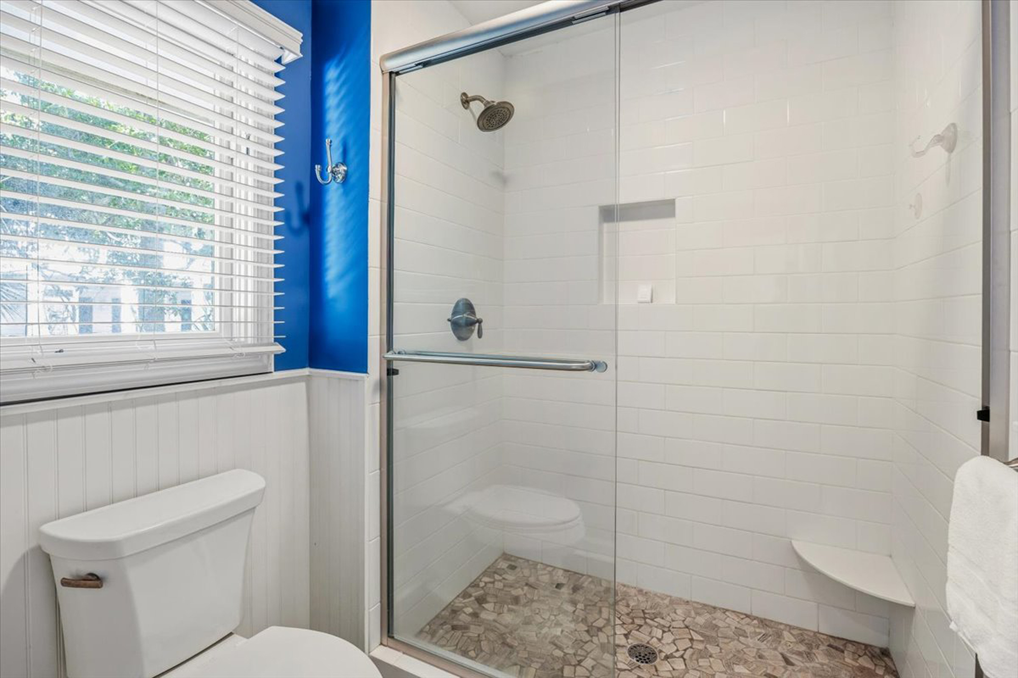 Deep blue walls meet classic white subway tile in this refreshing bathroom. A pebble stone shower floor adds an earthy texture underfoot, while daylight filters through the blinds to create a clean and airy morning atmosphere.