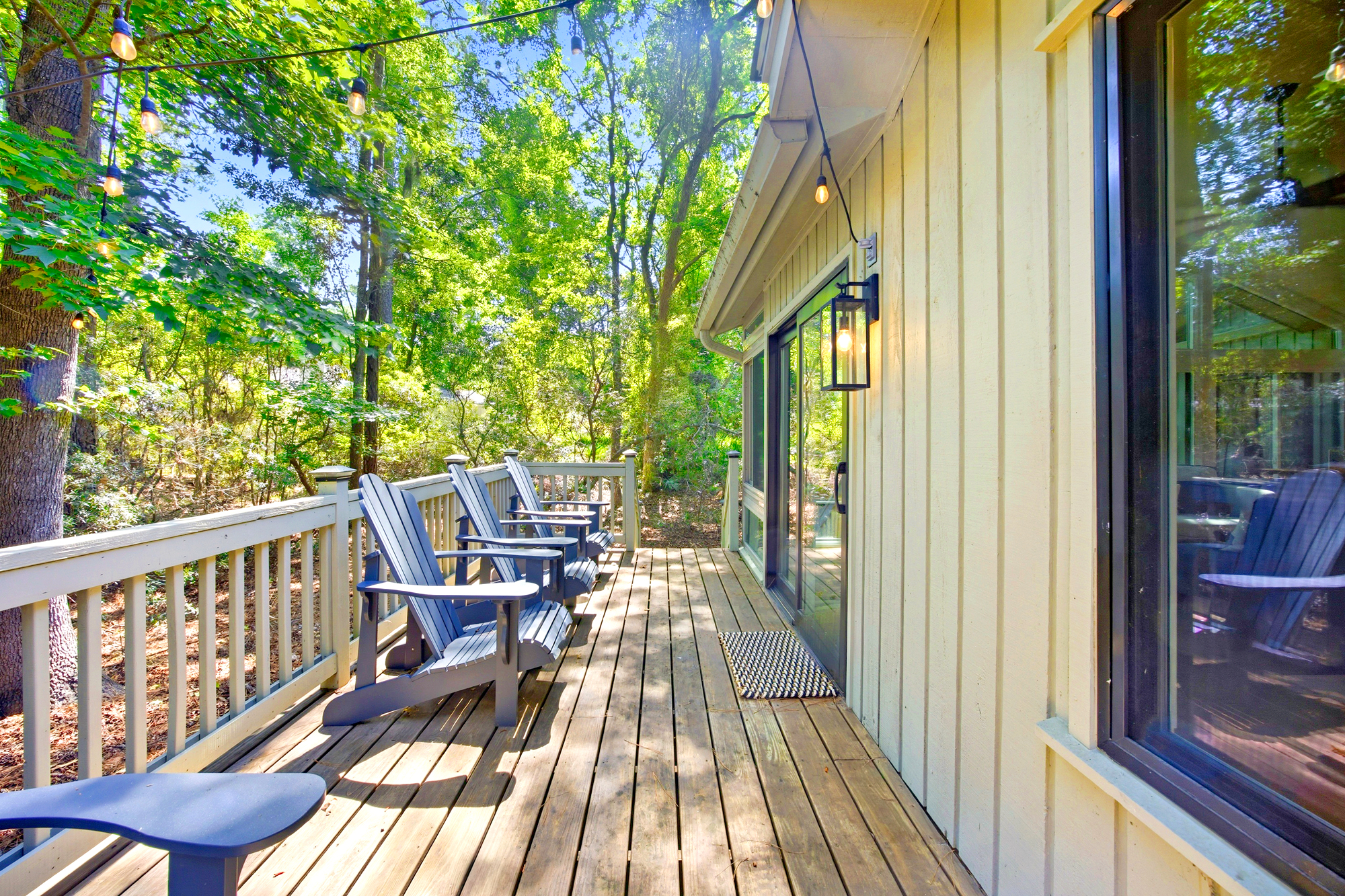 Adirondack chairs line this sun-drenched wooden deck, offering a quiet perch among the lush canopy of trees. Golden afternoon rays filter through the leaves, while overhead cafe lights promise a magical glow for your evening conversation.