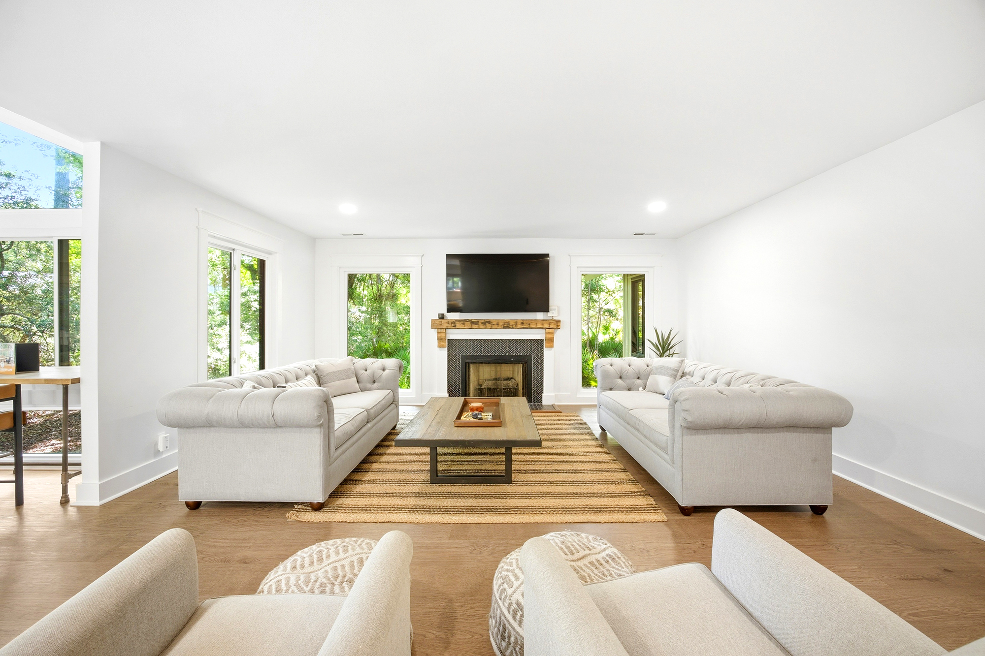 Plush tufted sofas face a rustic wood mantel and fireplace, creating a cozy hub for conversation. Large windows pull the garden's leafy greens into the bright, airy room, while a woven rug adds earthy texture to the golden timber floors.