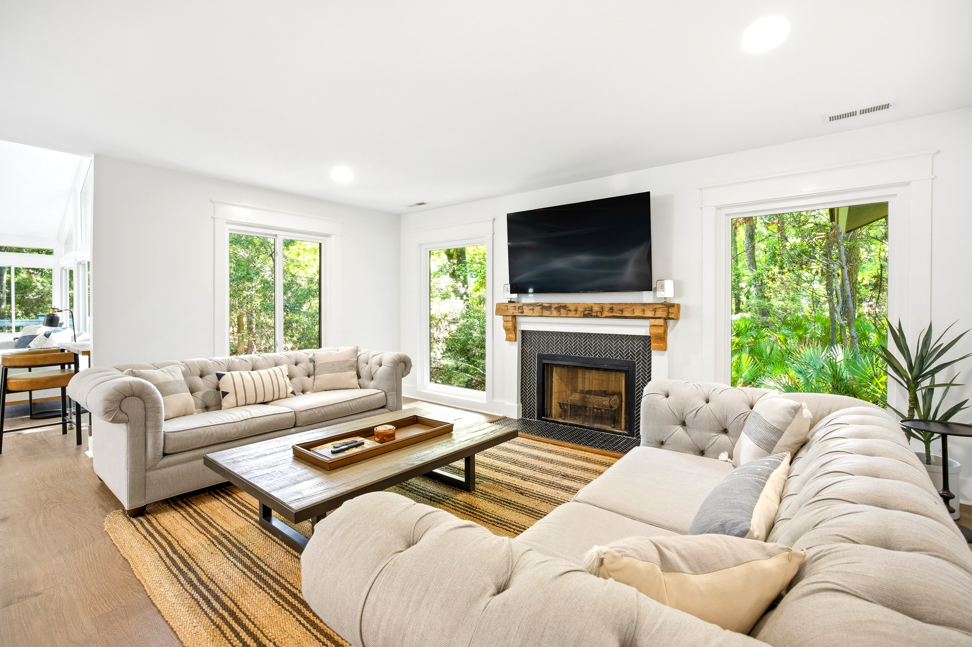 Soft linens and reclaimed wood accents create a grounded, airy lounge. Large windows draw in a bright afternoon glow, framing the lush greenery outside while the fireplace offers a cozy anchor for slow evenings at home.