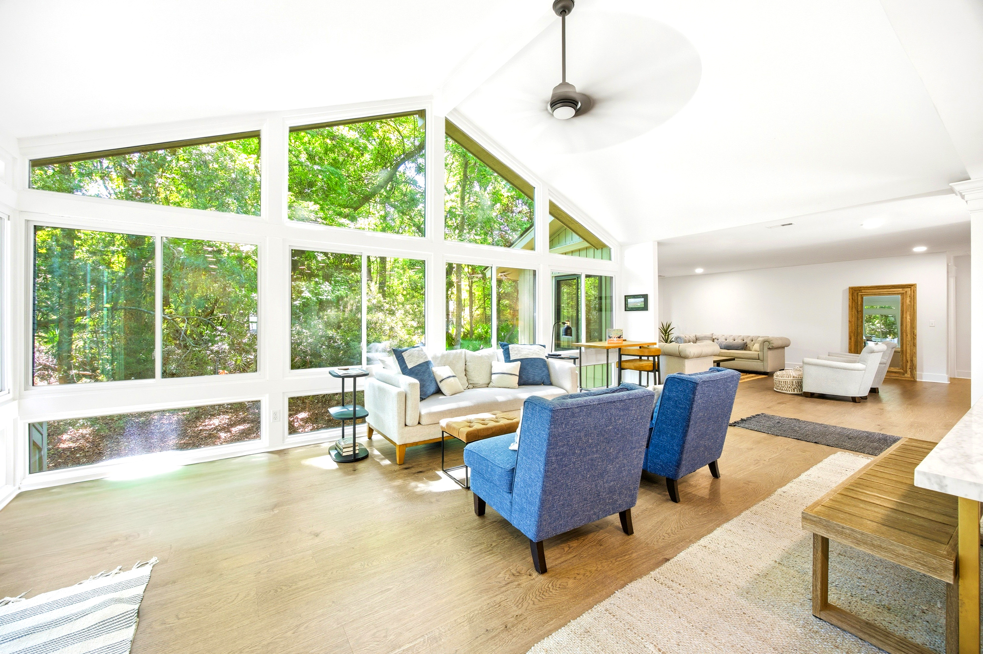 Floor-to-ceiling windows frame a lush canopy of green, drawing the outdoors into this airy, sun-drenched living room. Plush blue armchairs and oak floors create a warm, tactile space perfect for slow mornings and quiet conversation.