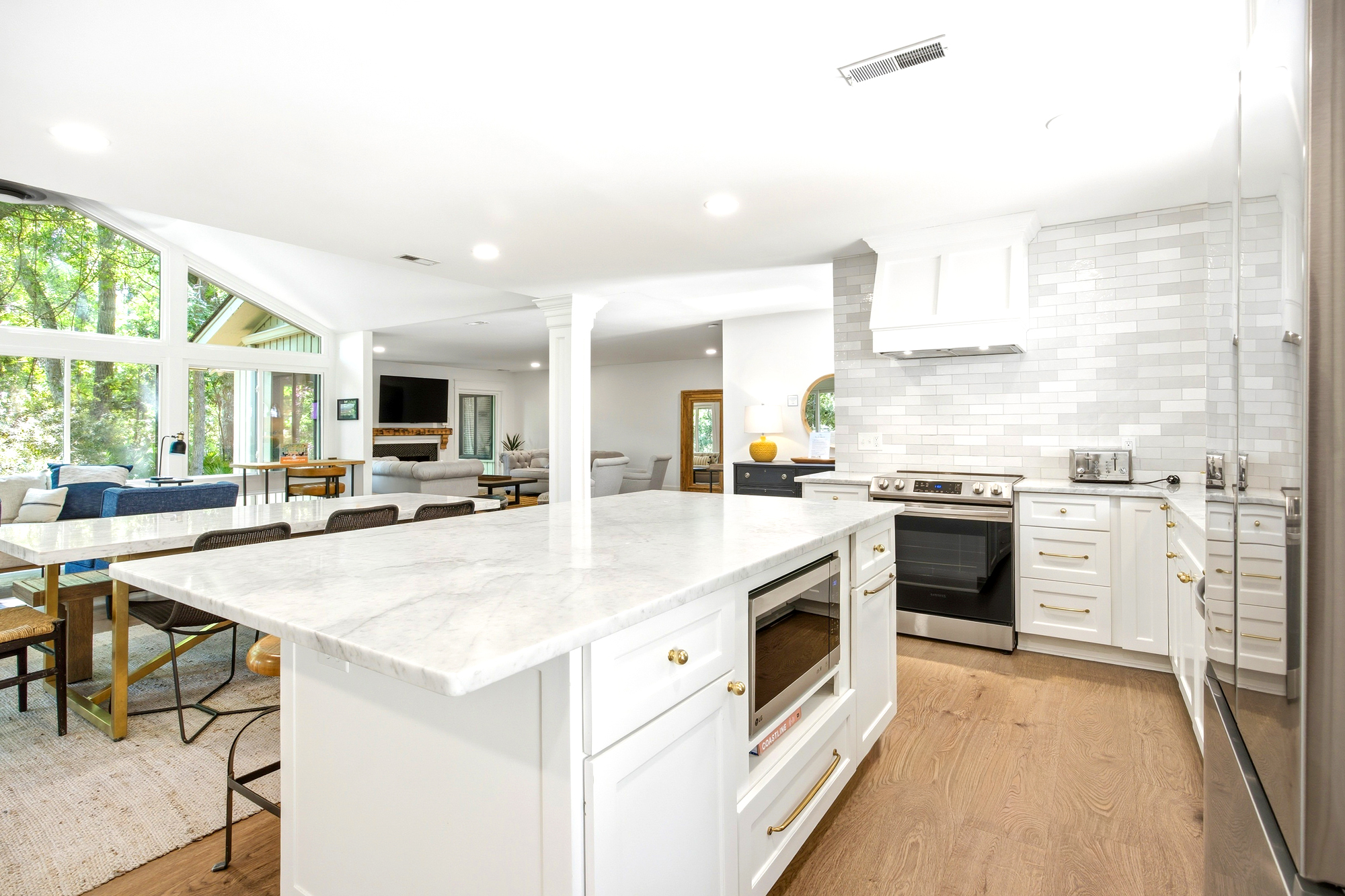 Marble countertops glisten under soft recessed lighting, providing a sleek surface for morning coffee. This airy kitchen flows into a bright living space where large windows frame peaceful views of the surrounding trees and greenery.