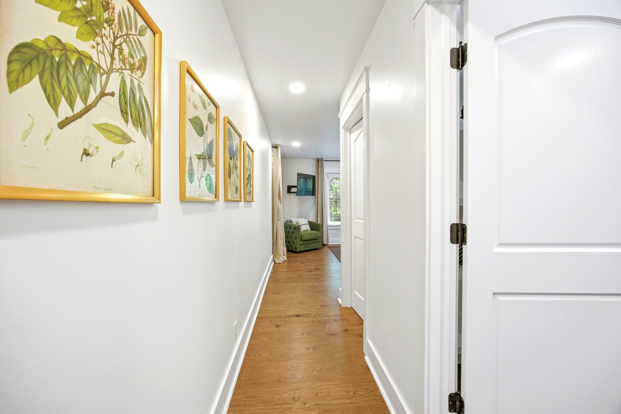 Botanical prints in golden frames line this airy hallway, leading your eyes toward a cozy reading nook. Pale walls and smooth wood planks catch the afternoon glow, creating a bright and peaceful passage through the home.