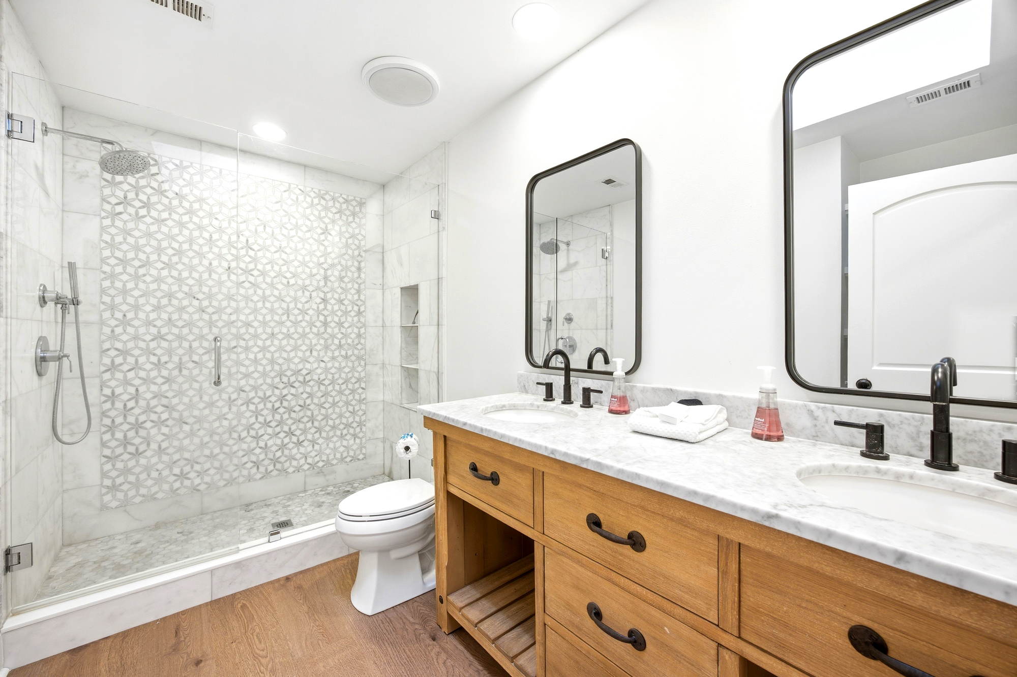 Oak cabinetry adds a rustic touch to this spa like suite, where intricate stone tiling and a spacious glass shower create a serene morning atmosphere. Bright, airy finishes pair with matte black hardware for a refined and refreshing vanity space.