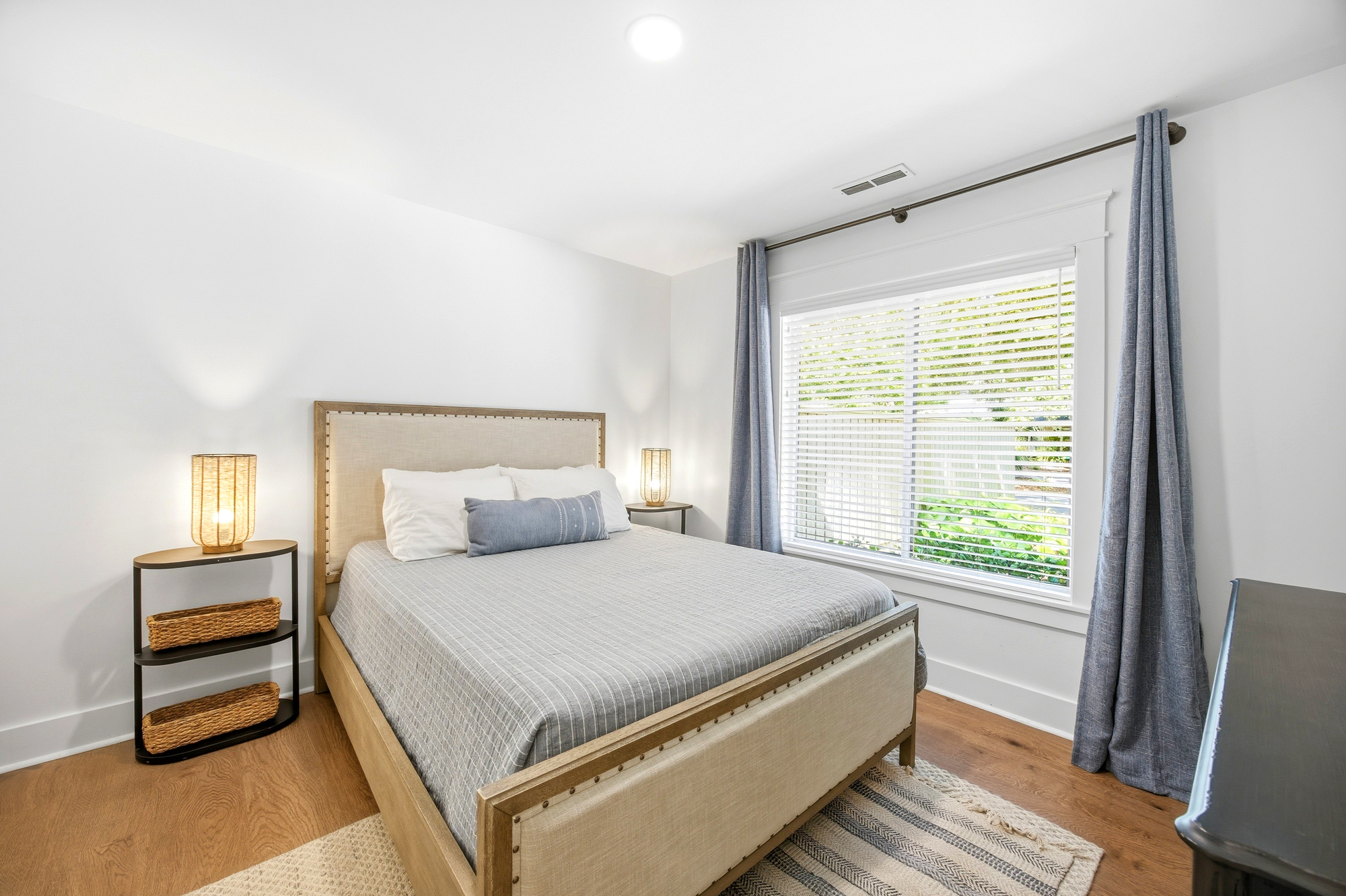 Blue denim drapes frame leafy garden views, casting a calm hum over this airy bedroom. Wicker accents and a studded fabric headboard add earthy textures to the sun-drenched space, creating a peaceful corner for quiet mornings.