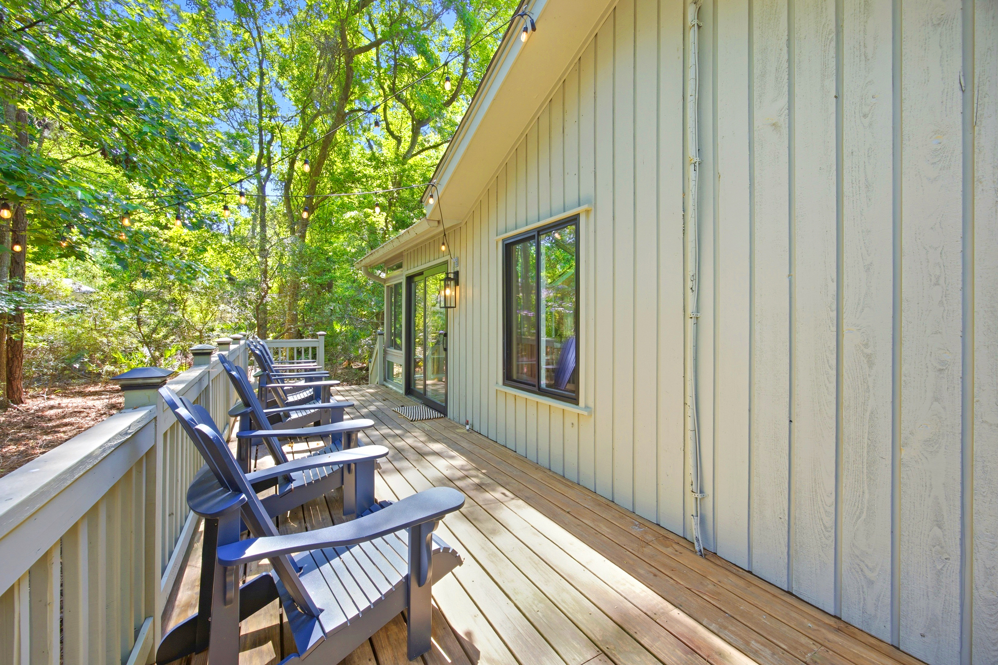 Weathered wood planks catch the afternoon glow on this expansive deck, where navy chairs wait under a canopy of twinkling bistro lights. Tall trees frame the scene, offering a peaceful screen of green for your quiet morning coffee or starlit chats.