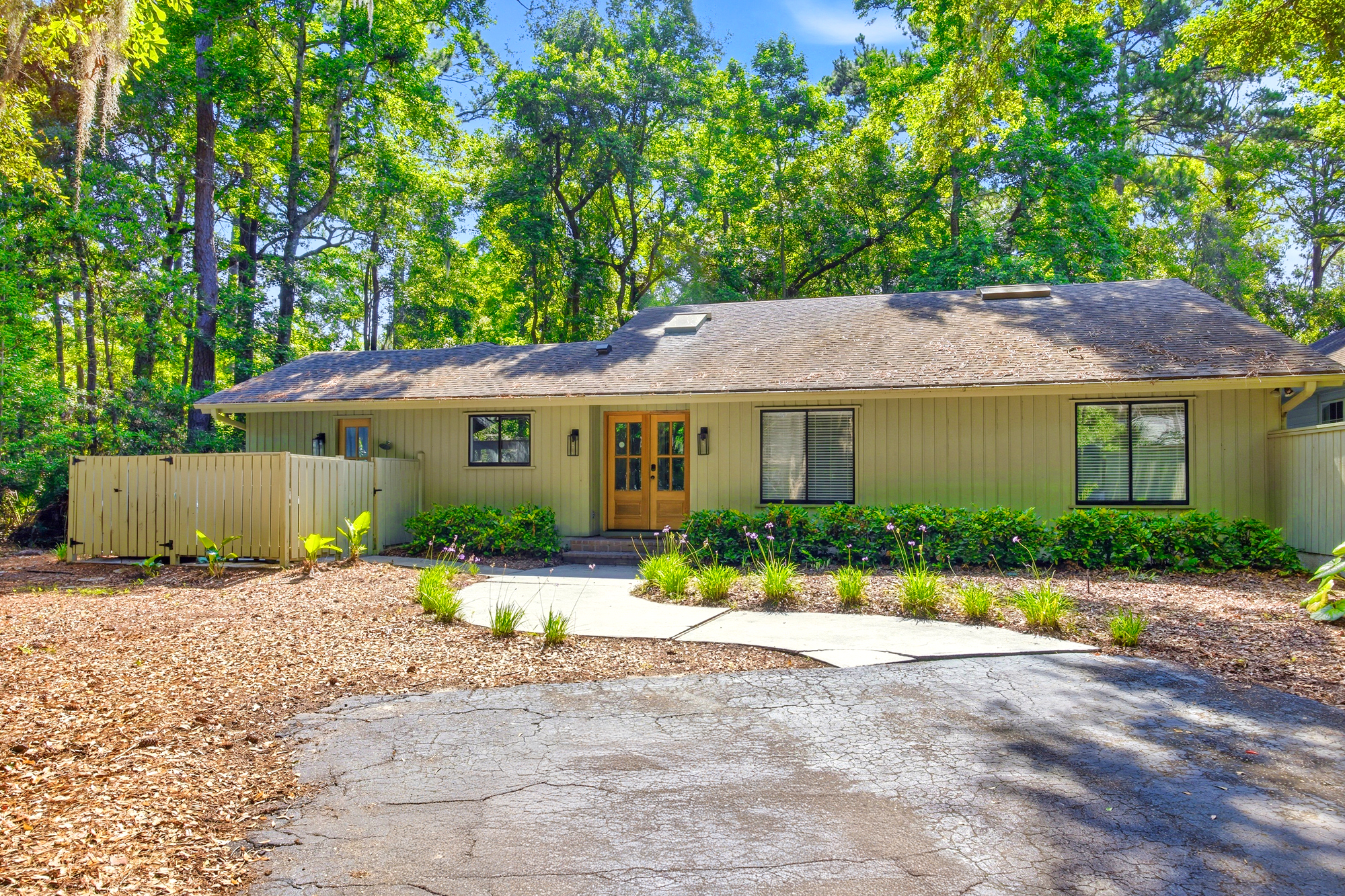 Tangled oaks and lush forest greens frame this serene cottage, where a winding path leads to inviting wooden double doors. Bright afternoon glow illuminates the olive siding and peaceful garden beds for a quiet, secluded arrival.