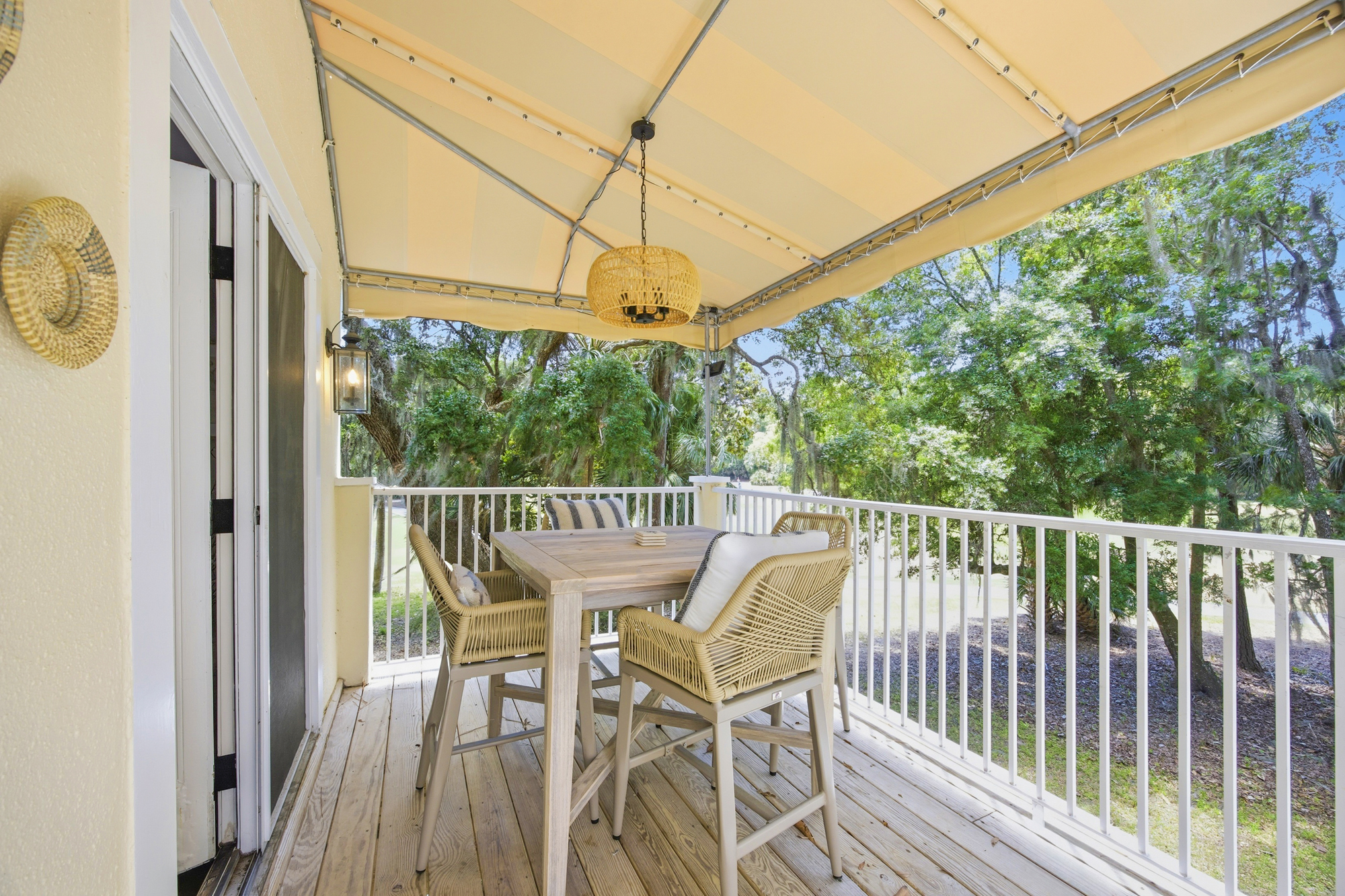 Braided wicker chairs and a pale wood table sit under a scalloped awning, creating a shaded nook for alfresco dining. Golden afternoon rays filter through the oak canopy, casting a calm, dappled glow across this breezy elevated terrace.