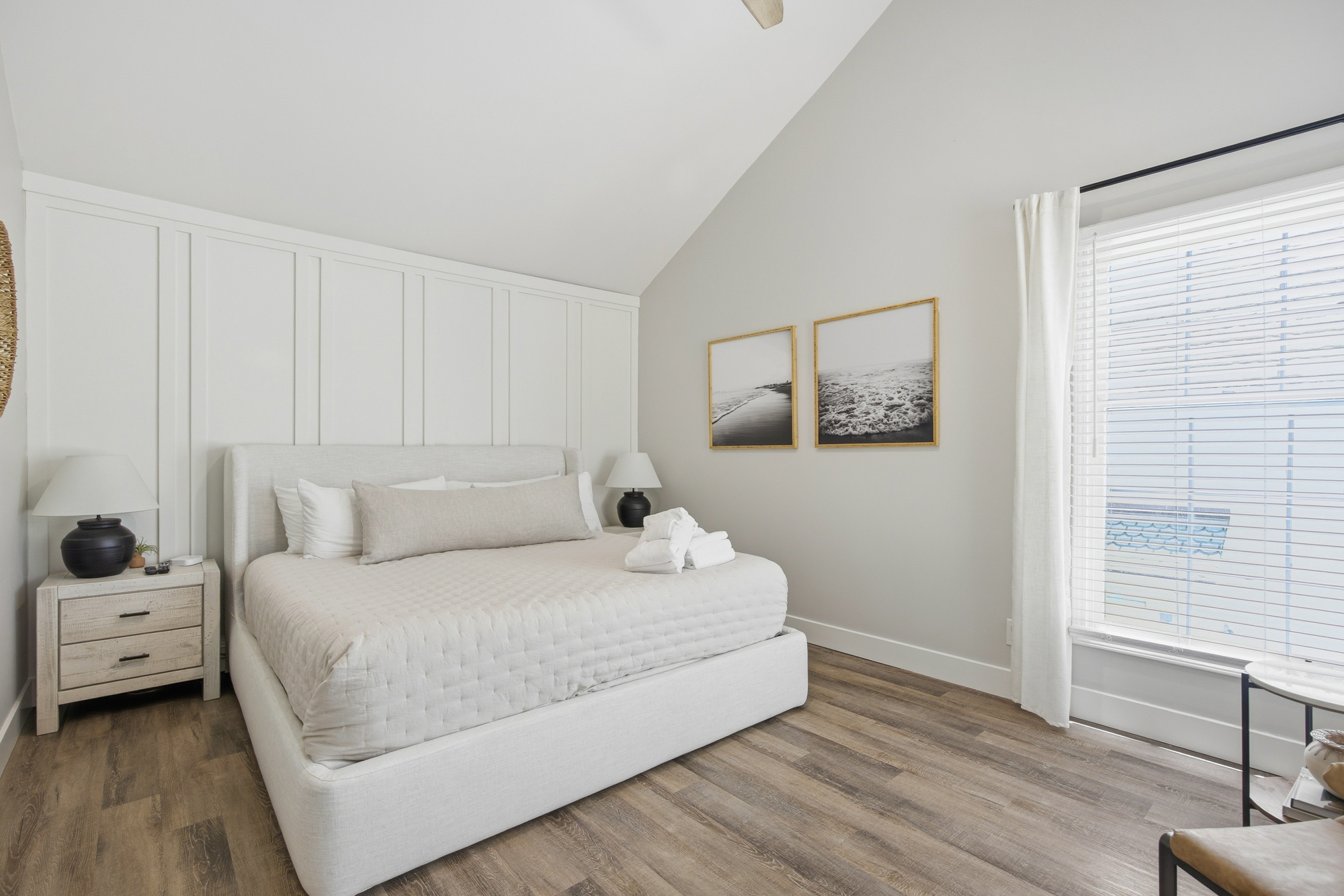 Creamy paneled walls and vaulted ceilings create a sense of airy calm in this primary suite. Soft textures meet warm wood floors, while daybreak filters through the window to illuminate the plush linens and coastal art.