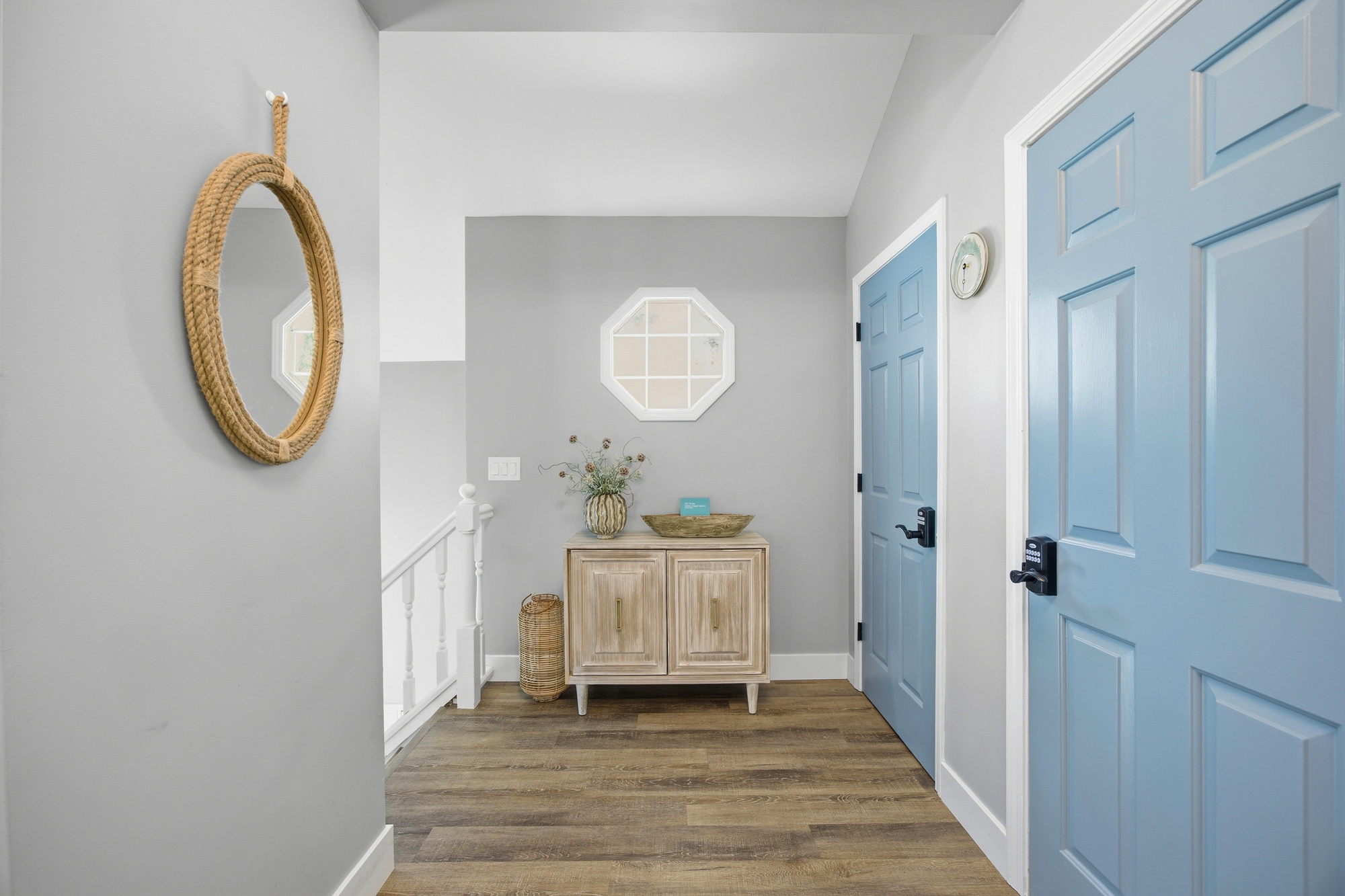 Sky-blue doors add a cheerful pop of color to this breezy hallway, where a rope-framed mirror and pale wood console create a serene arrival. Gentle overhead light reflects off the smooth gray walls for a clean and calming transition.