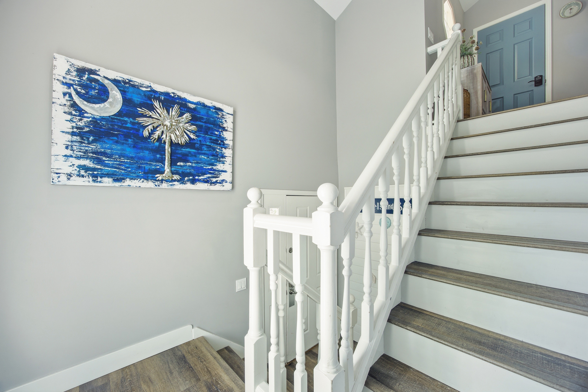 Vibrant local art pops against smooth dove-gray walls, leading your eye up a gracious white staircase. Deep wood treads and a wash of airy daylight create a cheerful, open transition between the cozy levels of this seaside home.