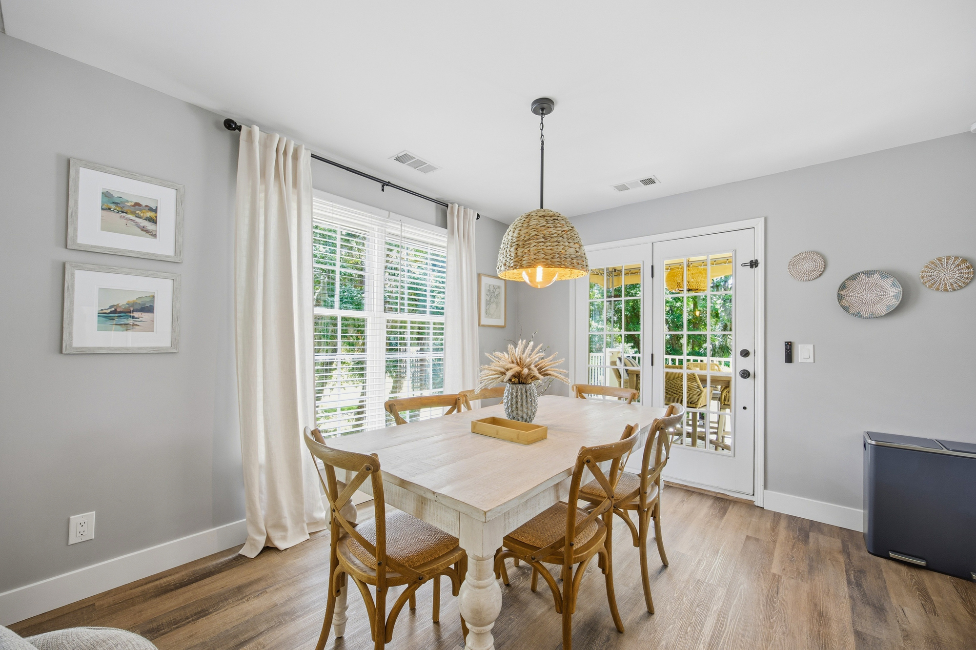Sun-drenched windows and garden views frame this airy dining nook, where hand-woven textures and warm wood tones create a gentle setting for morning coffee. A farmhouse table sits under the glow of a wicker pendant, perfect for slow brunch.
