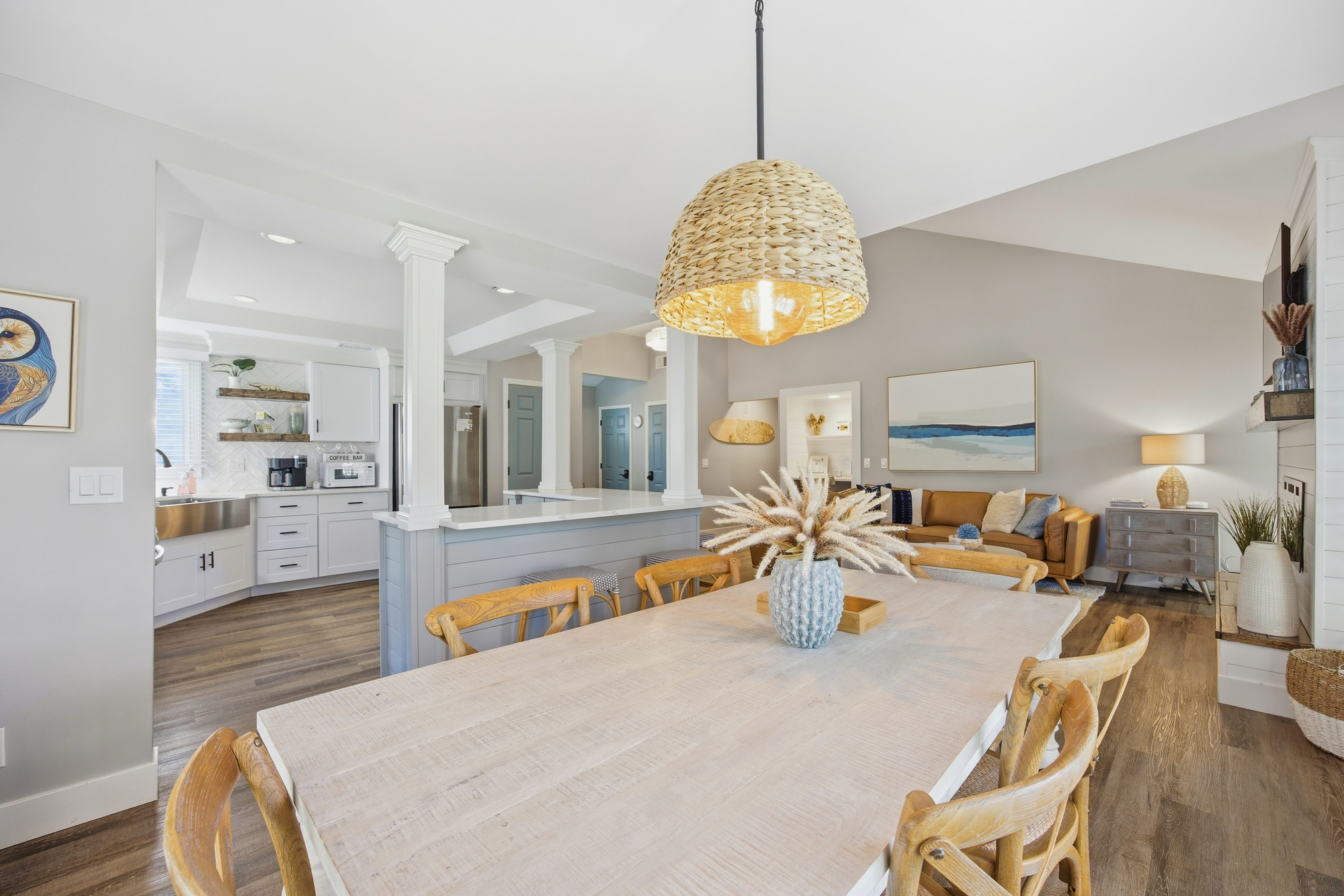 Weathered wood textures and an airy open floor plan create a coastal flow from the dining table to the modern kitchen. A statement wicker pendant casts a golden glow over the long table, perfect for slow mornings and shared meals.