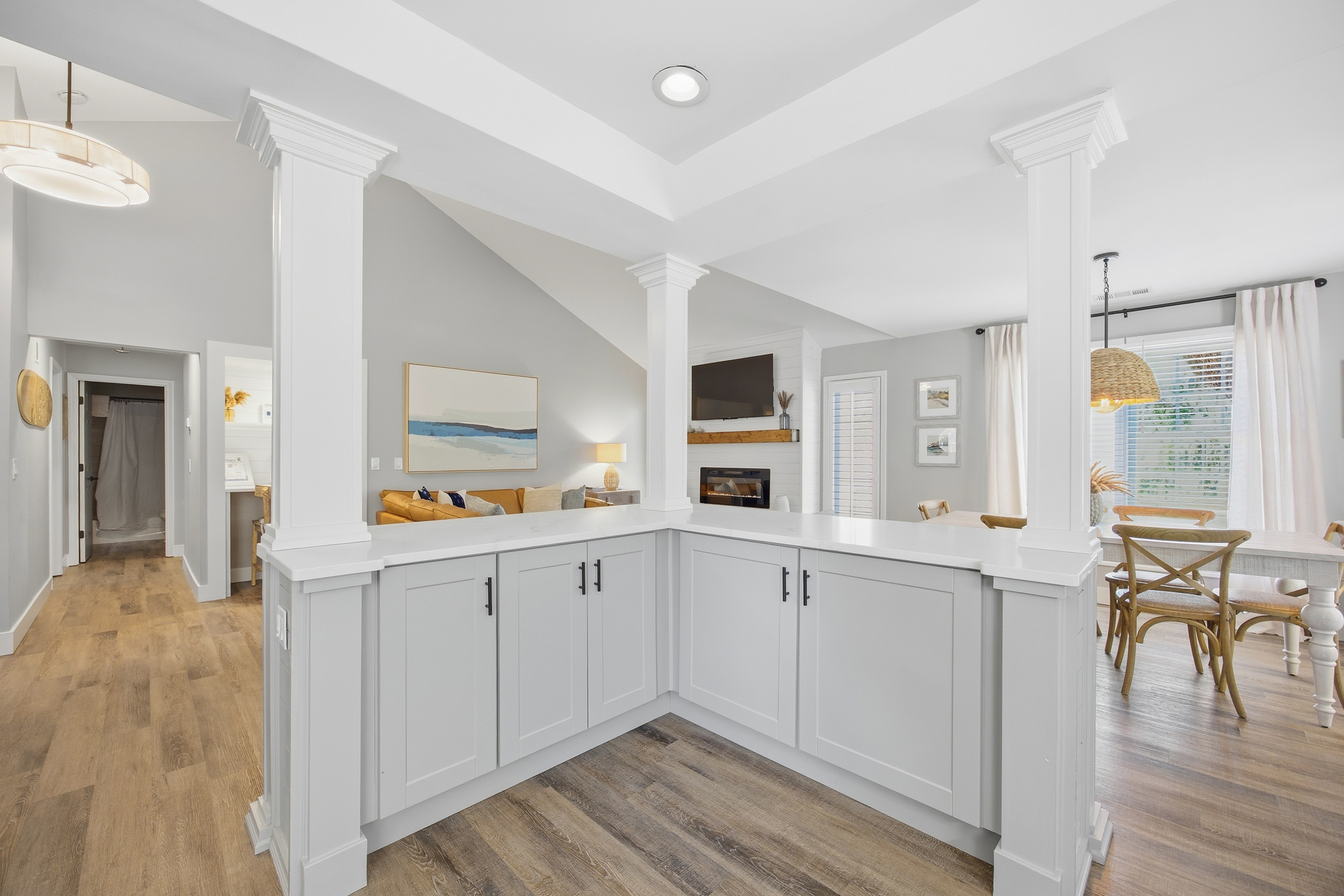 White pillars frame this airy open floor plan, where honey-toned wood floors lead your eye from the sleek kitchen counters to a sun-drenched dining nook. Warm textures and bright, gallery-style walls create a rhythmic, peaceful flow.