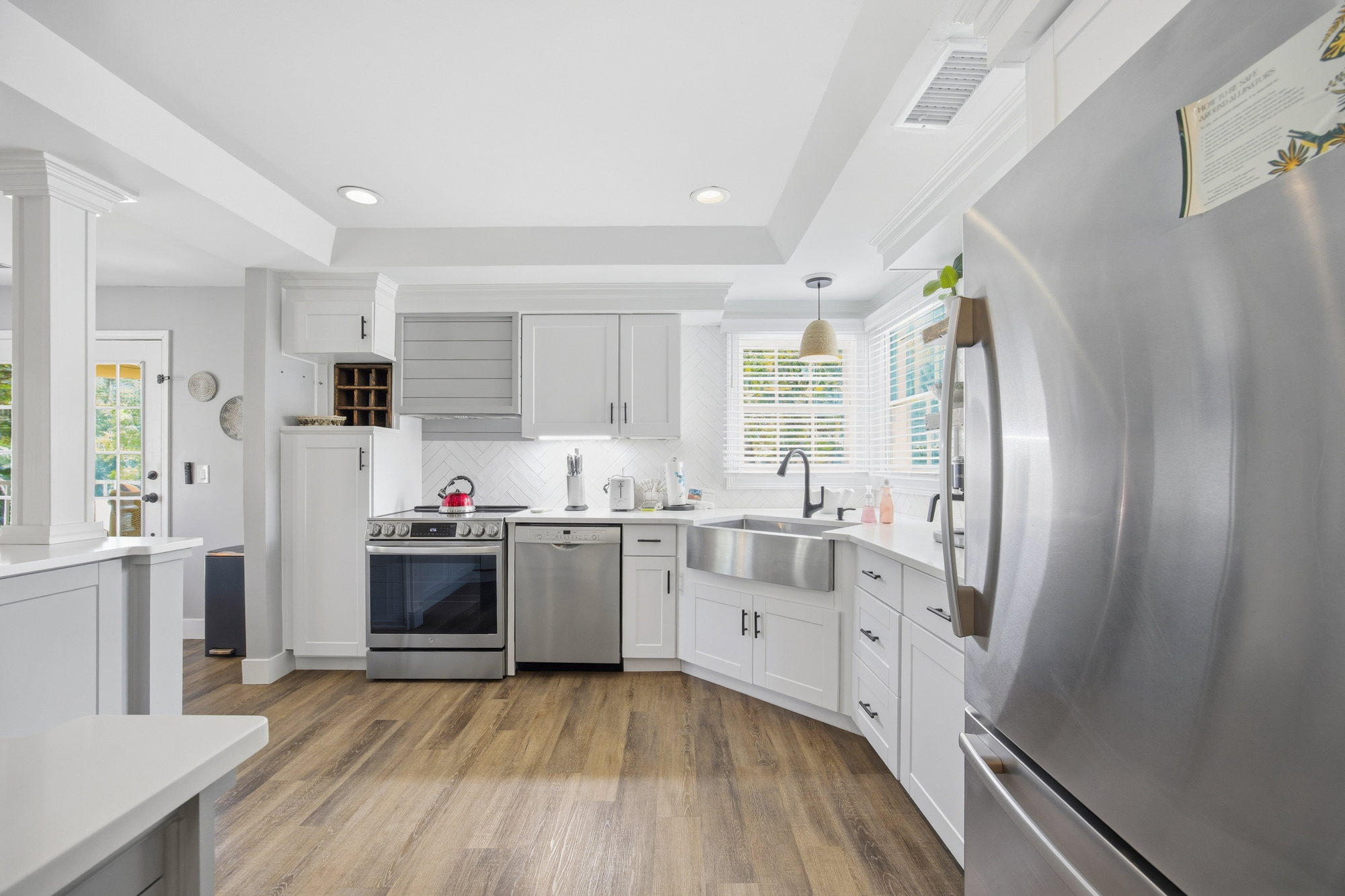 Stainless steel appliances and a deep farmhouse sink anchor this airy kitchen, where bright cabinetry meets warm wood plank flooring. Natural light pours over the herringbone tile backsplash, creating a cheerful space for morning coffee.