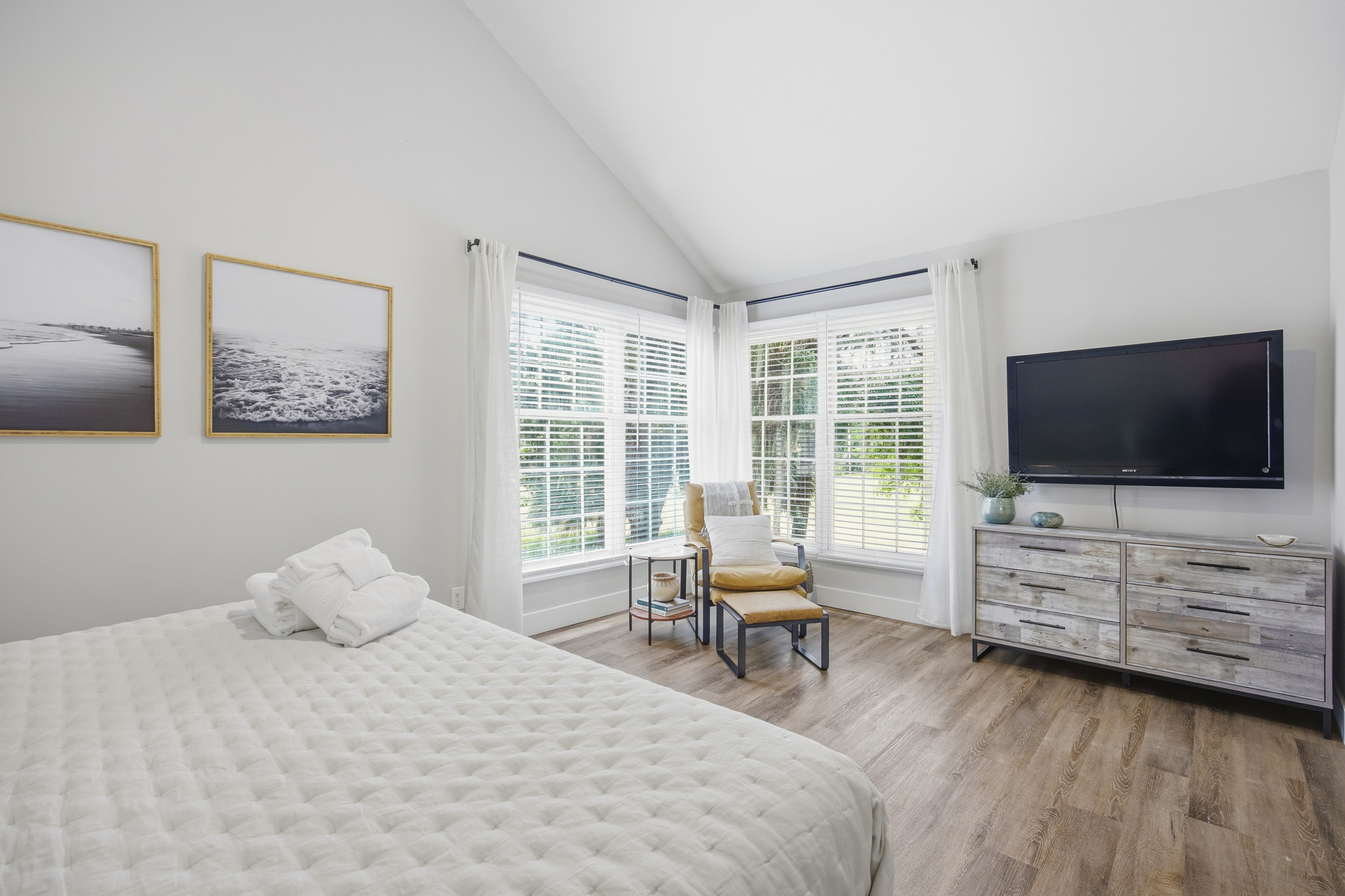 High ceilings and crisp white linens create a sense of airy calm in this sun-drenched bedroom. Weathered wood textures and a cozy reading nook by the window offer a serene spot to rest after a day of coastal adventures.