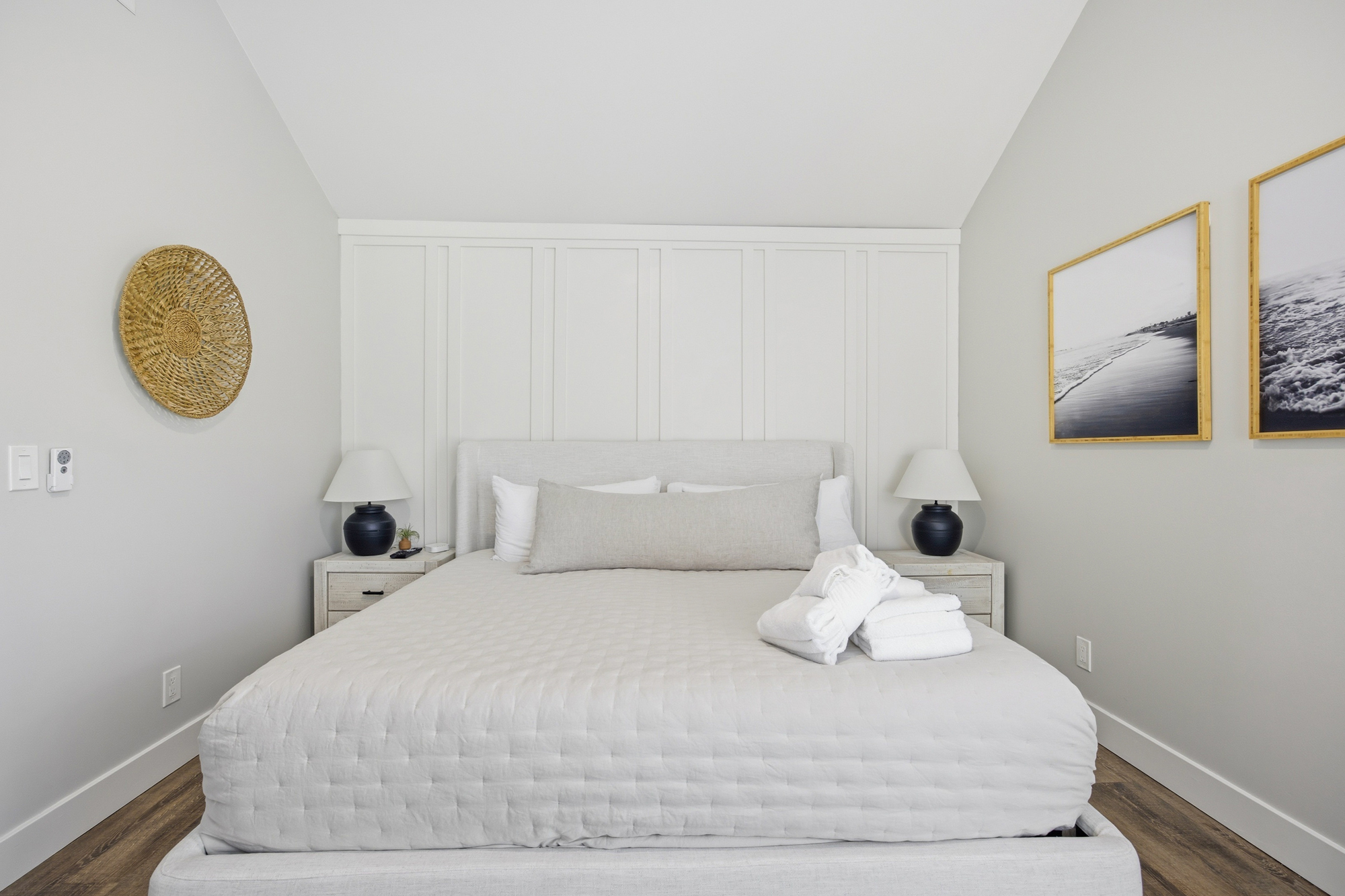 Crisp linens and textured pillows rest against a paneled feature wall, creating a serene sanctuary for sleep. Natural textures and coastal art bring a sense of calm to this bright, airy bedroom.