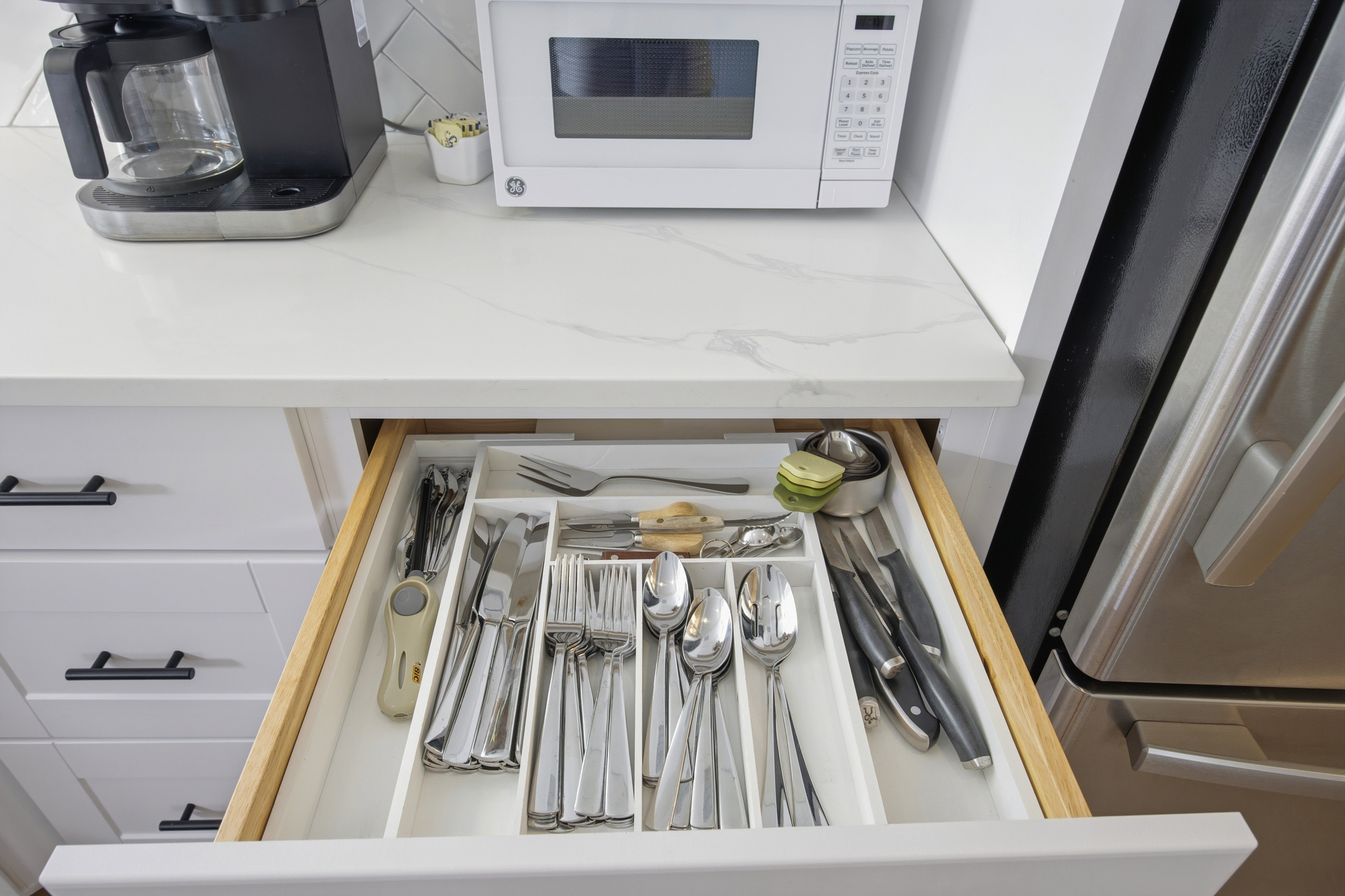 Gleaming silverware rests in an organized drawer, ready for your next home-cooked meal. Marbled stone counters and bright white finishes create a clean, modern atmosphere for easy mornings in the kitchen.