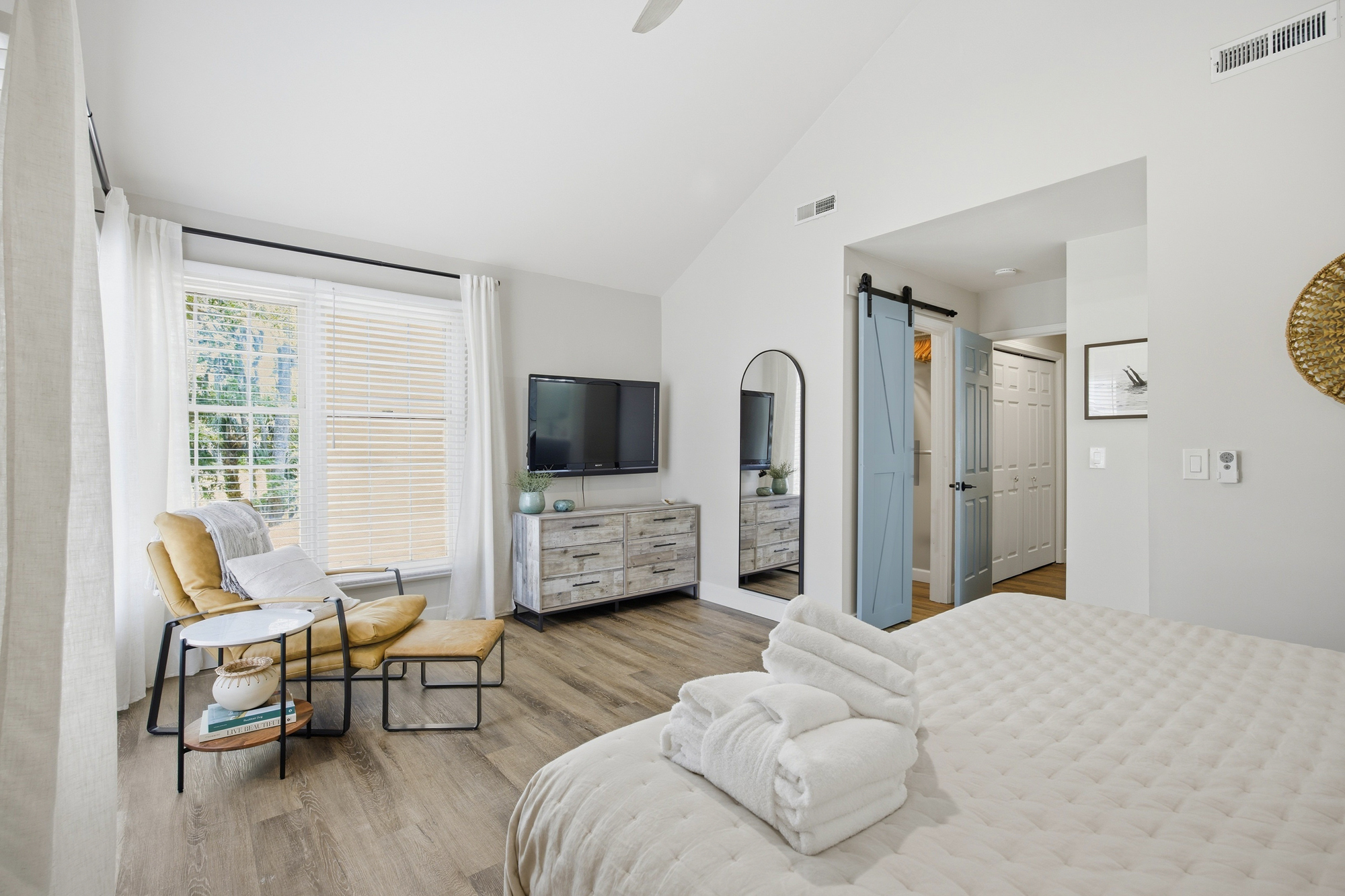 Vaulted ceilings and soft morning rays create an airy sanctuary, where a honey-toned leather chair invites you to linger. Weathered wood textures and a pop of dusty blue add a calm, coastal character to this sun-drenched master suite.