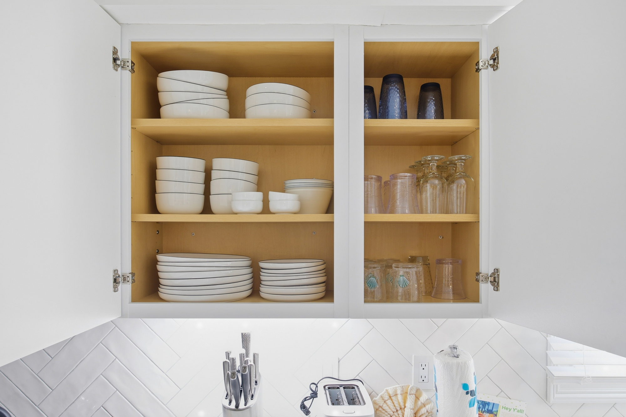 Tidy oak shelves house an array of naval blue glassware and ceramic dishes, all organized for your next seaside meal. Bright under-cabinet lighting glints off the fresh white subway tile, creating a cheerful corner for morning prep.
