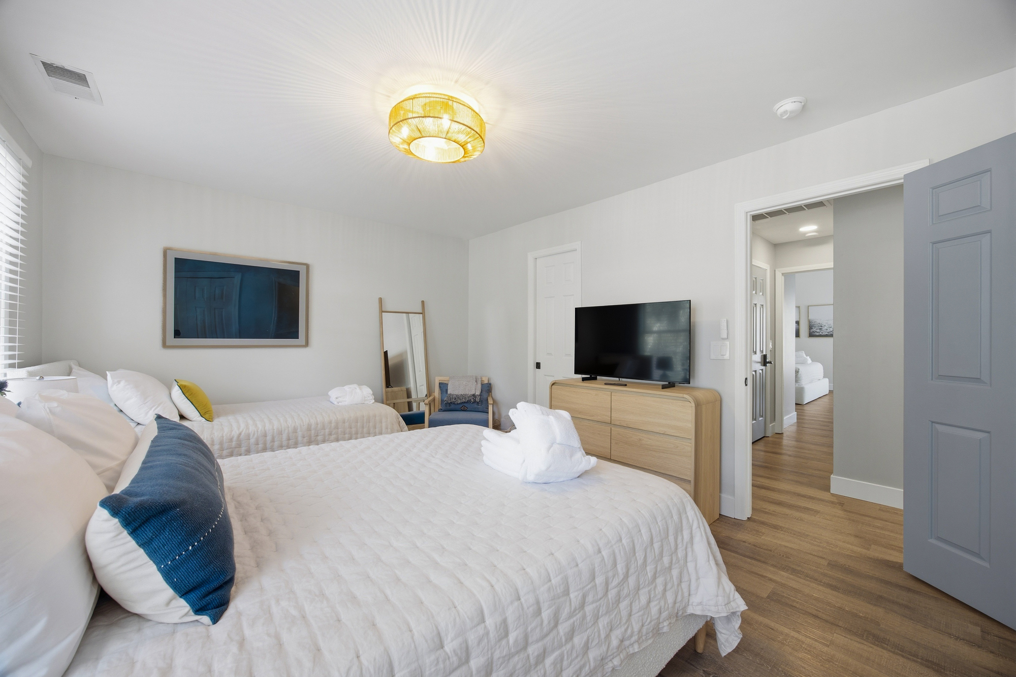 Woven textures and sea-blue accents create a peaceful atmosphere in this bright bedroom. A honey-toned light fixture casts a warm radiance over the plush linens and smooth wood floors, making it a cozy spot for a quiet evening.