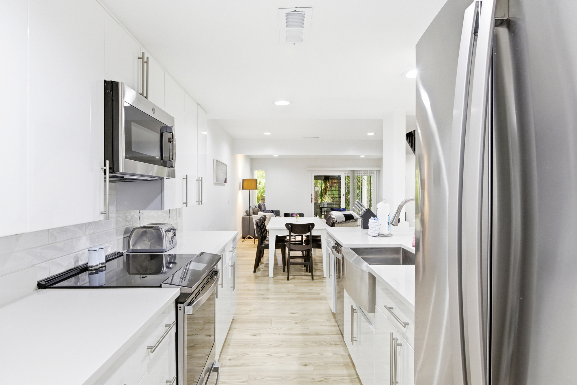 Stainless steel appliances and snowy surfaces create a culinary space that feels airy and organized. Soft overhead glow illuminates the open galley, leading your eyes toward the shared dining area and the leafy views beyond.