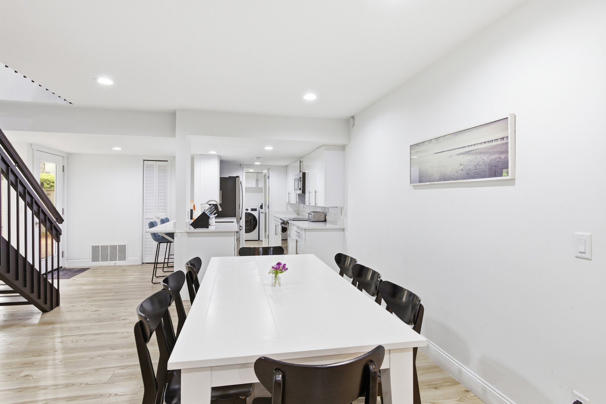 Bright white surfaces and dark wood accents create a balanced, airy dining space perfect for group meals. The open layout flows seamlessly from the kitchen, illuminated by soft overhead lighting and a glimpse of the outdoors.