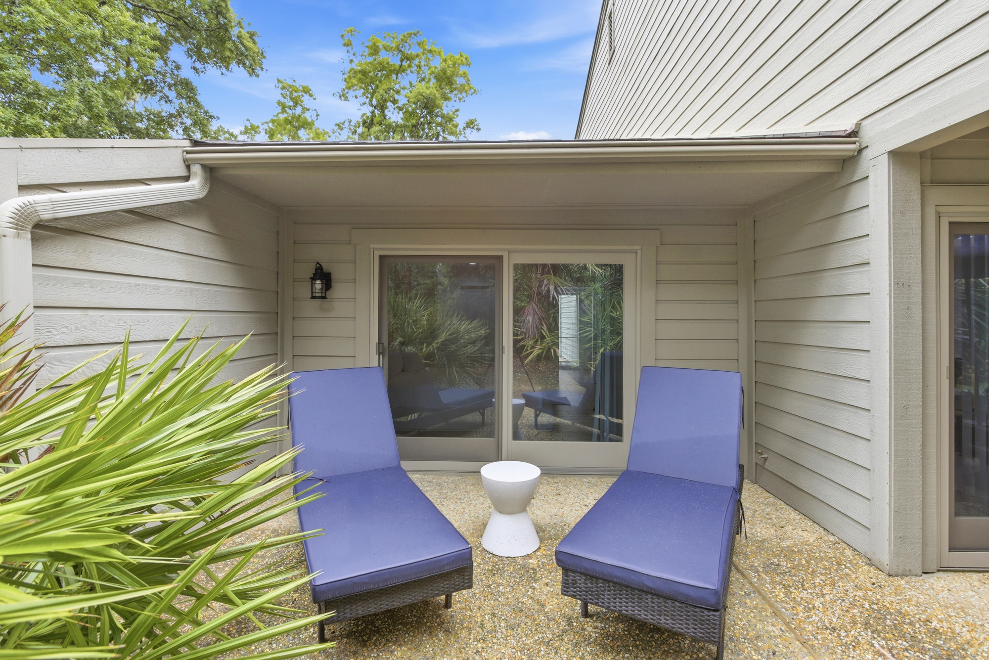 Indigo outdoor loungers wait on a pebbled patio under the shade of a wide overhang. Verdant palm leaves frame this quiet nook, creating a private corner to sip a cool drink and watch the afternoon sky.