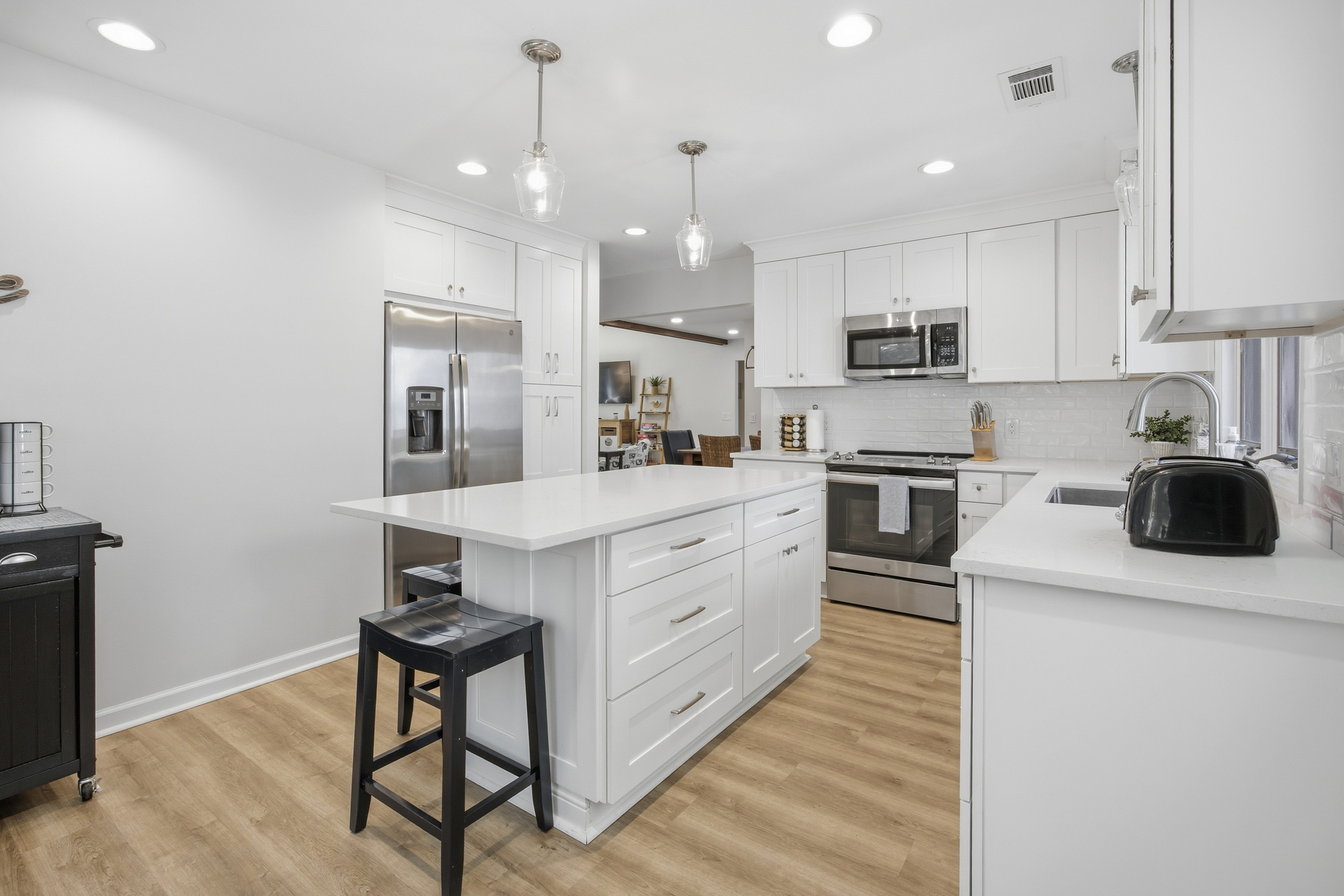 Pristine white cabinetry and quartz counters create a gleaming culinary space, grounded by oak toned flooring. Pendant lights cast a gentle radiance over the central island, making this modern kitchen the heart of your morning routine.