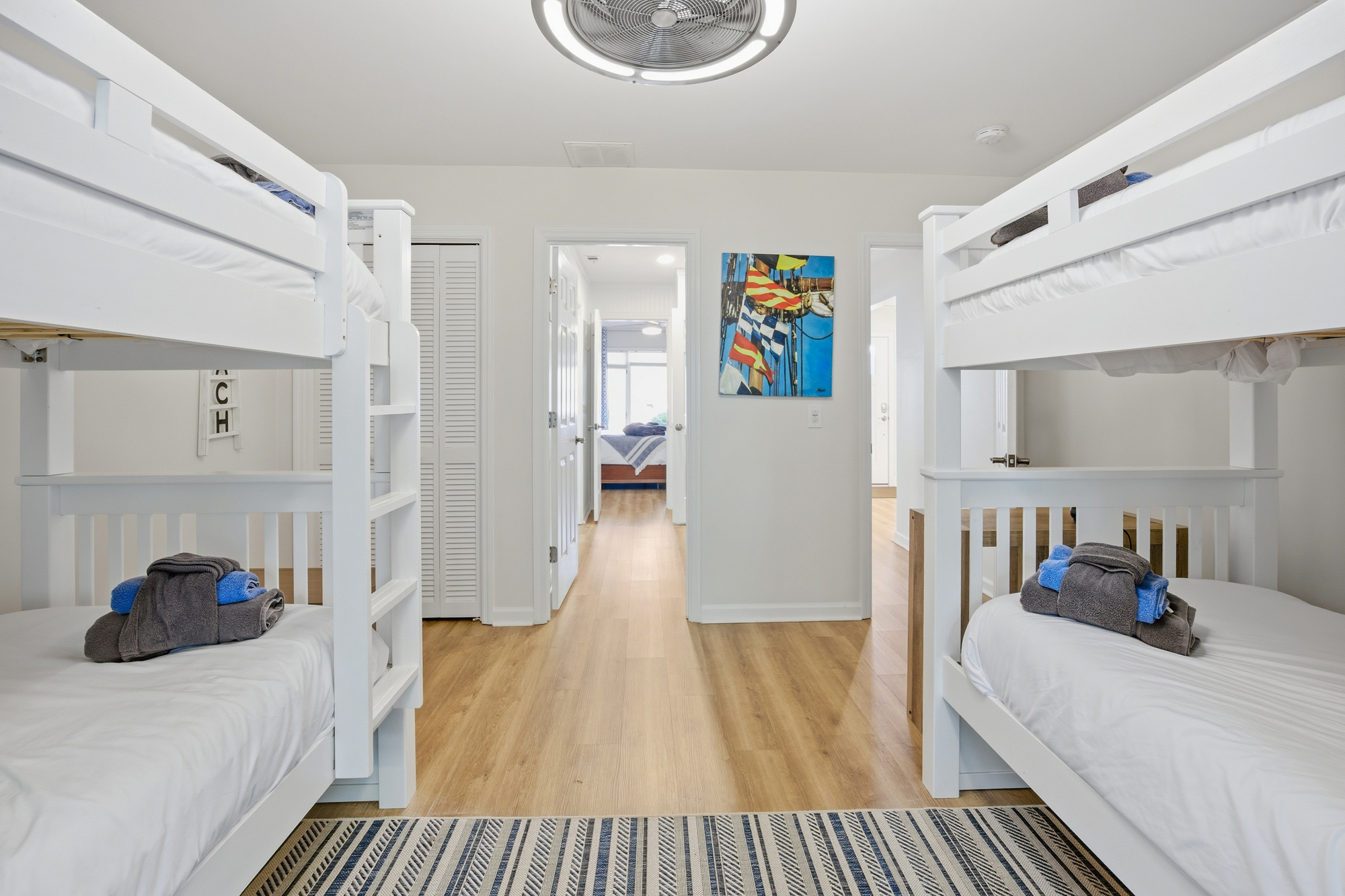Smooth white bunk beds offer a playful yet serene spot for guests to rest, featuring fresh linens and nautical accents. Golden oak planks lead your eye through the airy hallway, where coastal artwork adds a splash of color to this cheerful space.